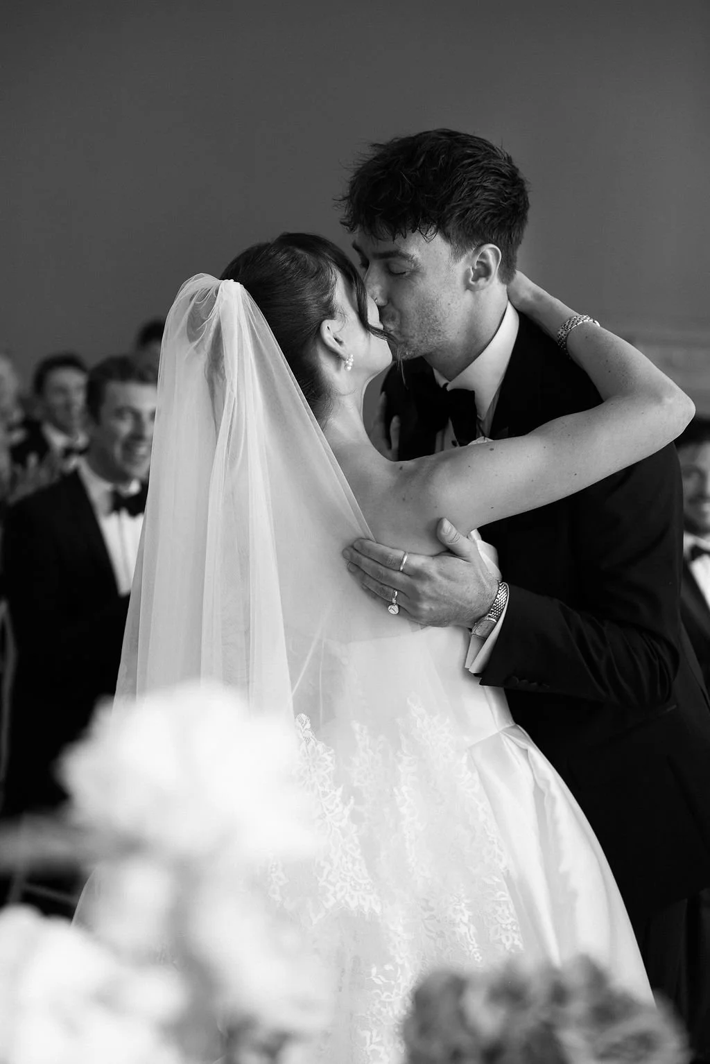 Black and white photo of a bride and groom kissing during their wedding ceremony, with guests in formal attire visible in the background.