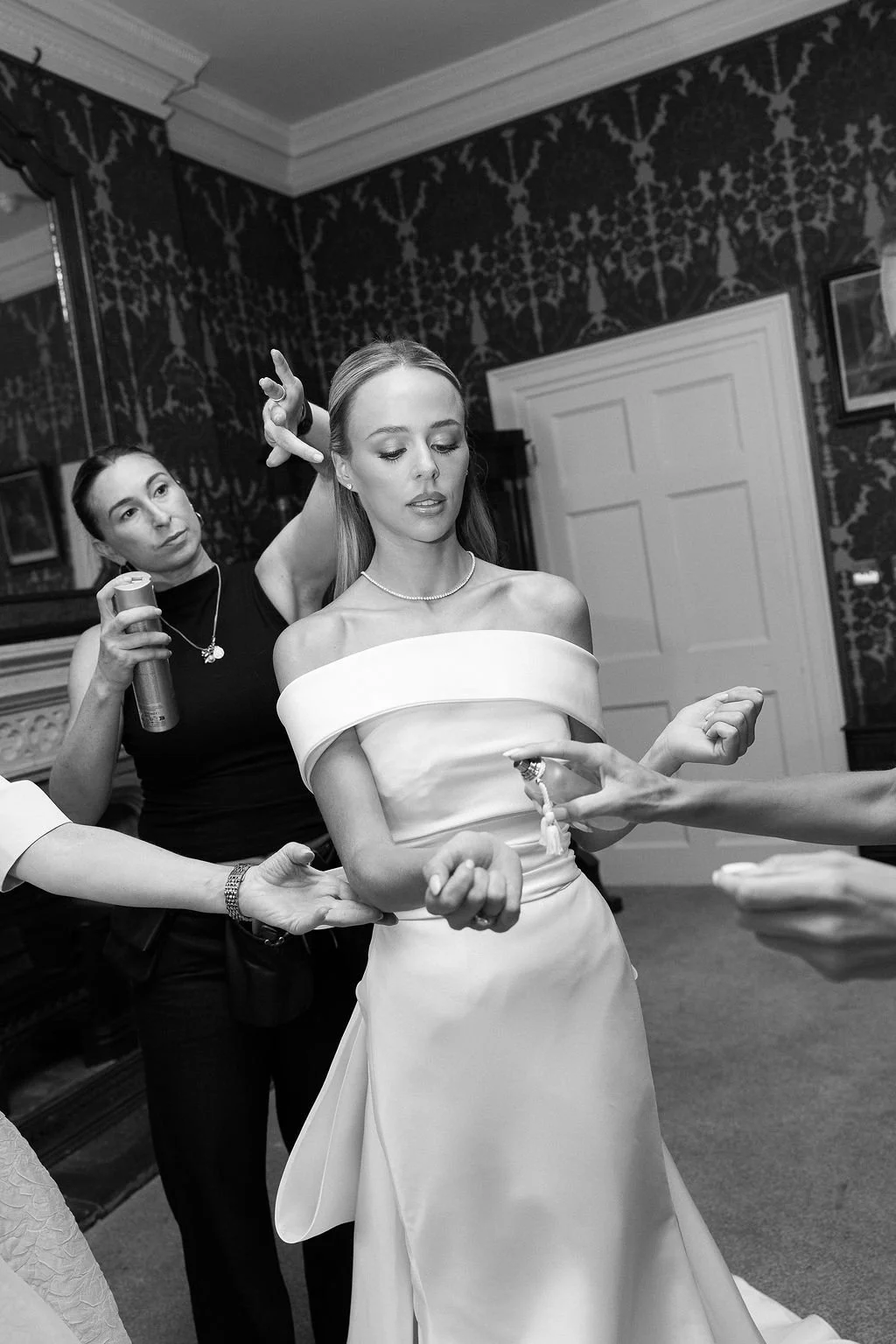 Woman in an off-the-shoulder satin wedding dress being assisted with jewelry, surrounded by people preparing her for a wedding.