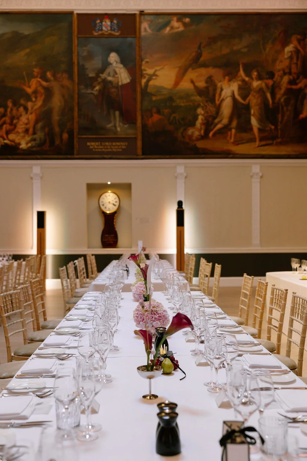 Elegant dining table set for a formal event in a grand room with artwork and a clock on the wall.