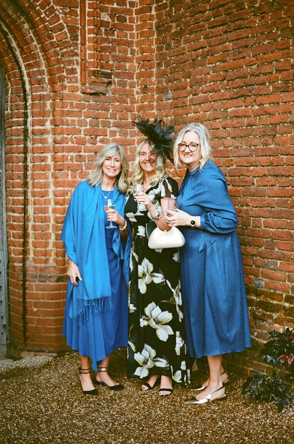 Three women dressed elegantly, standing together, smiling, and holding glasses of wine in front of a brick wall.