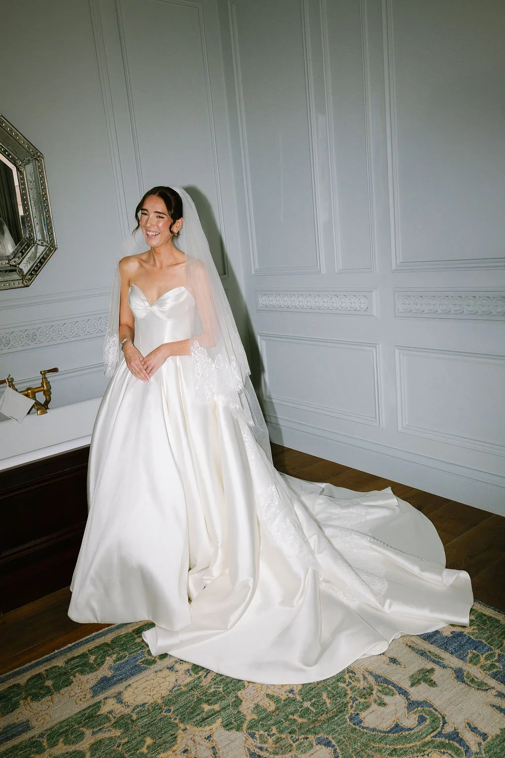 A smiling bride in a white wedding gown with a long train, standing in a room with white paneled walls and a large mirror. She is wearing a veil and jewelry, and appears happy.