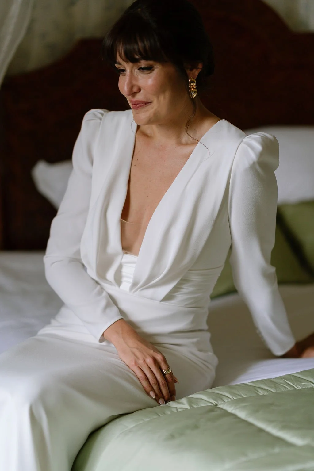 A woman with dark hair in a white dress, sitting on a bed with green bedding in a warmly lit room.
