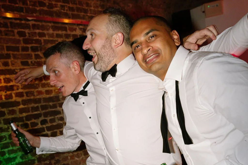 Three men in white shirts and black bow ties at a party, smiling and posing together in front of a brick wall, some holding drinks.