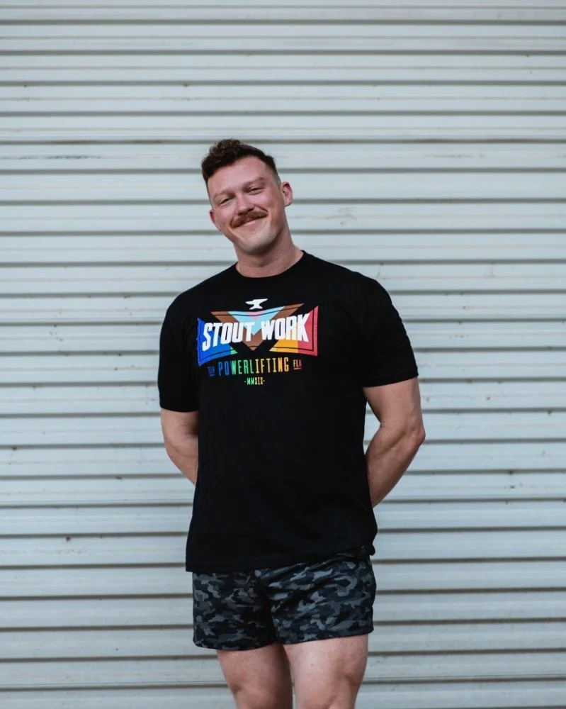 A man with short brown hair, a mustache, wearing a black t-shirt with colorful text, and camouflage shorts, standing outdoors in front of a gray metal garage door, smiling with his hands behind his back.