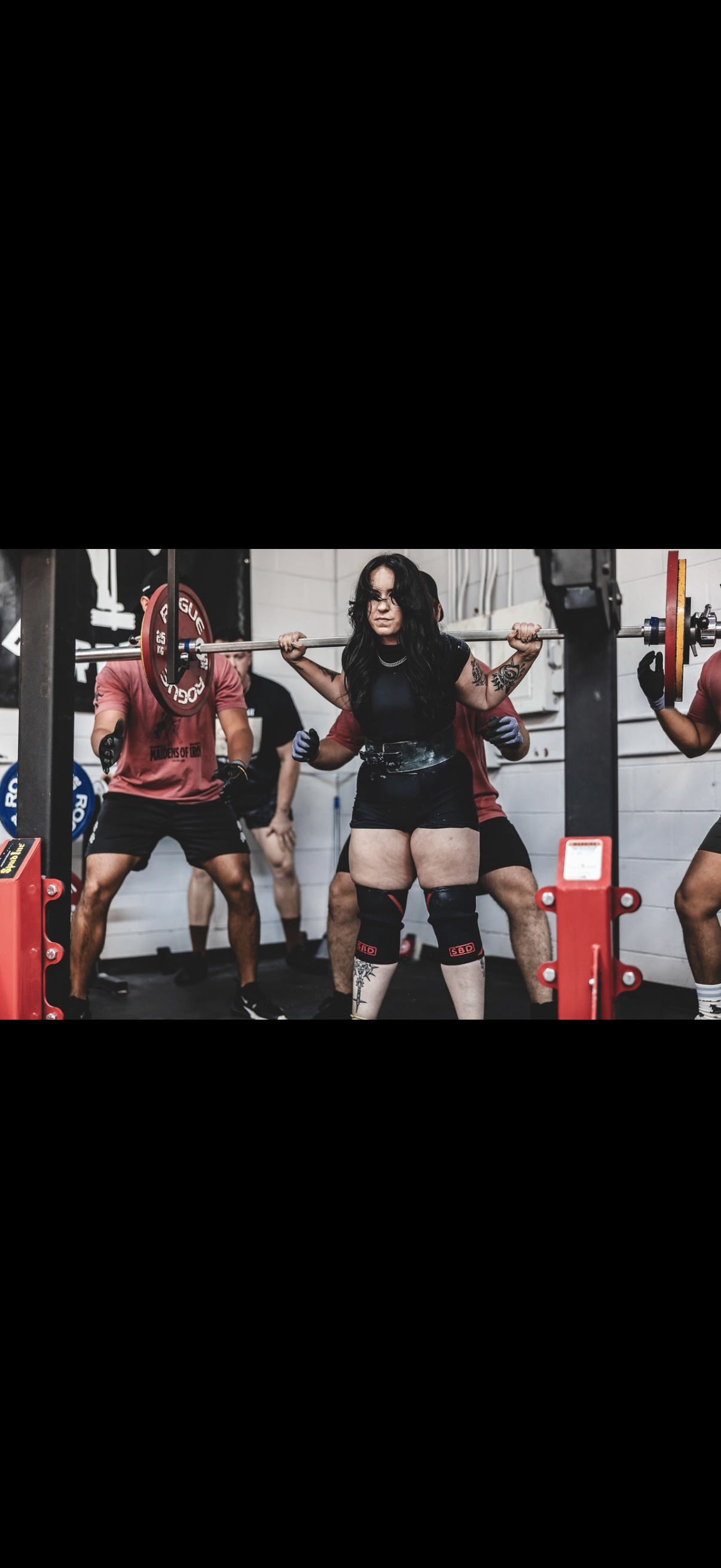 Woman lifting a barbell during a weightlifting workout, surrounded by several individuals in a gym setting.