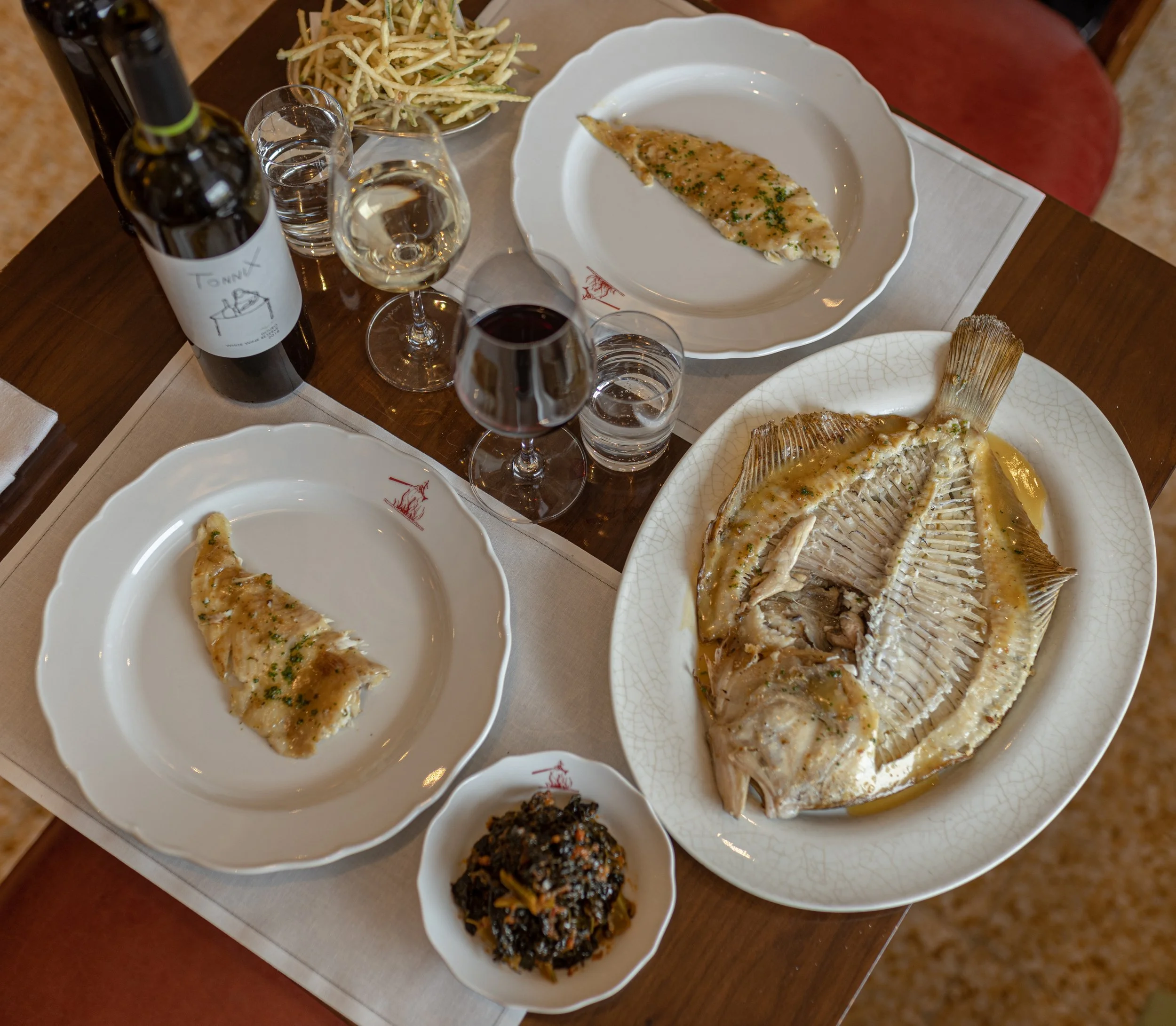 A dining table with plates of grilled fish garnished with herbs, bowls of side dishes including fried vegetable sticks, a piece of grilled fish on a plate, a bottle of red wine, and glasses of red and white wine.
