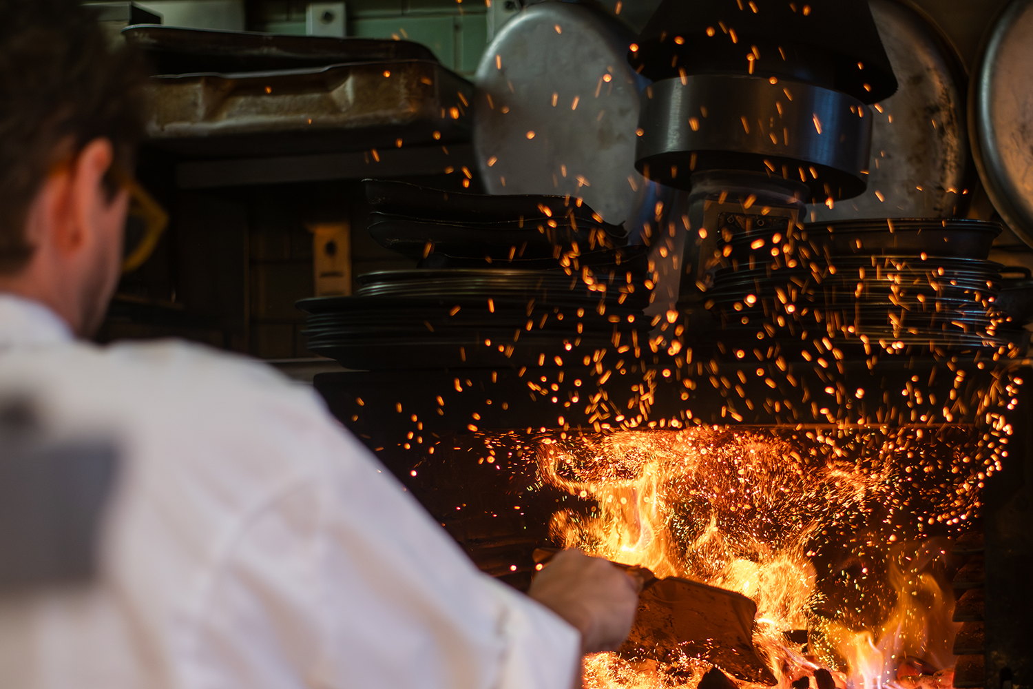 A chef in a kitchen is working with fire and sparks flying as they handle a piece of wood or food over an open flame, with stacked plates and cooking utensils in the background.