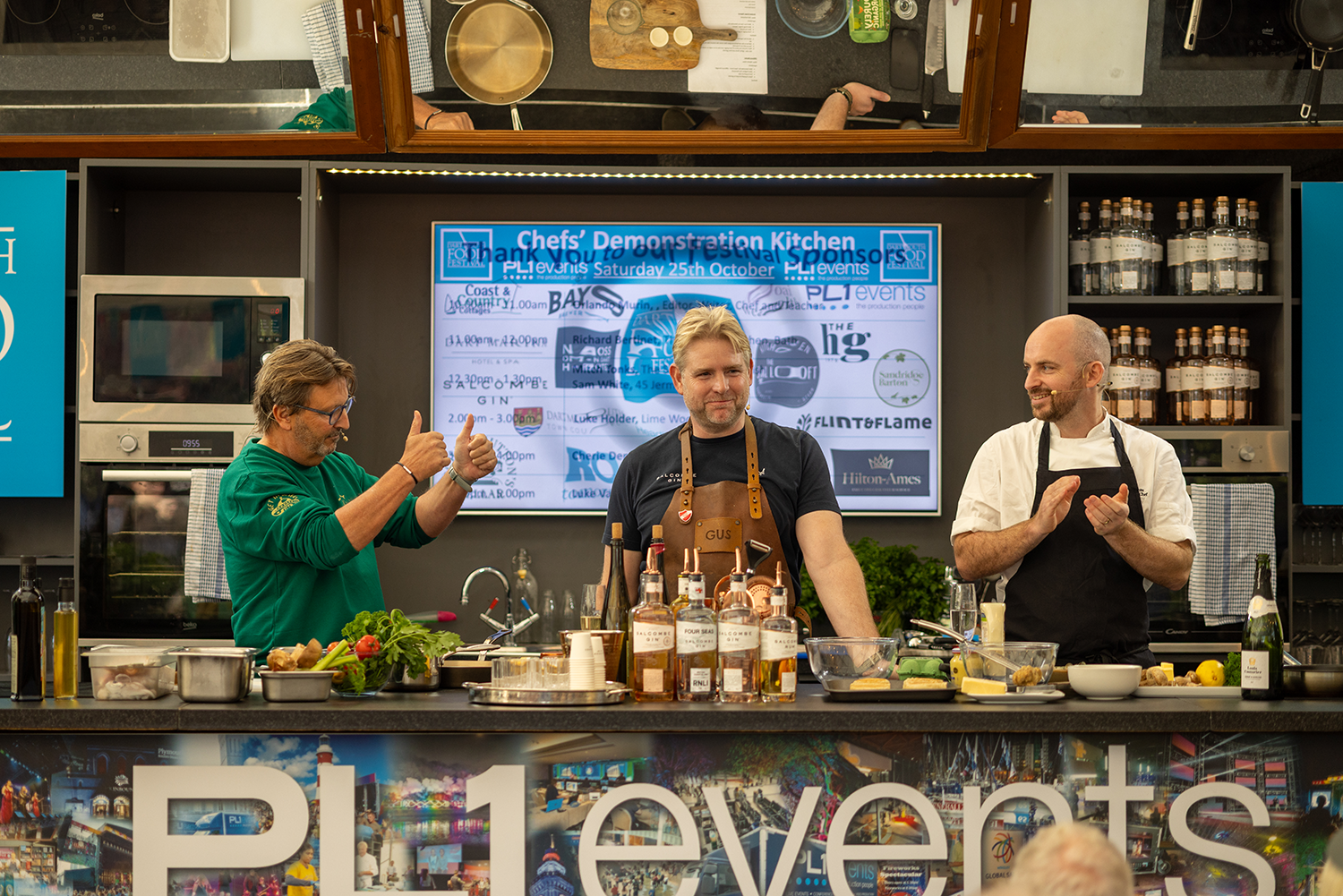 Three men standing behind a kitchen counter during a cooking demonstration in a chef's demonstration kitchen. They are smiling, clapping, and giving a thumbs up. The counter has cooking ingredients and bottles of alcohol. A large screen in the backgr