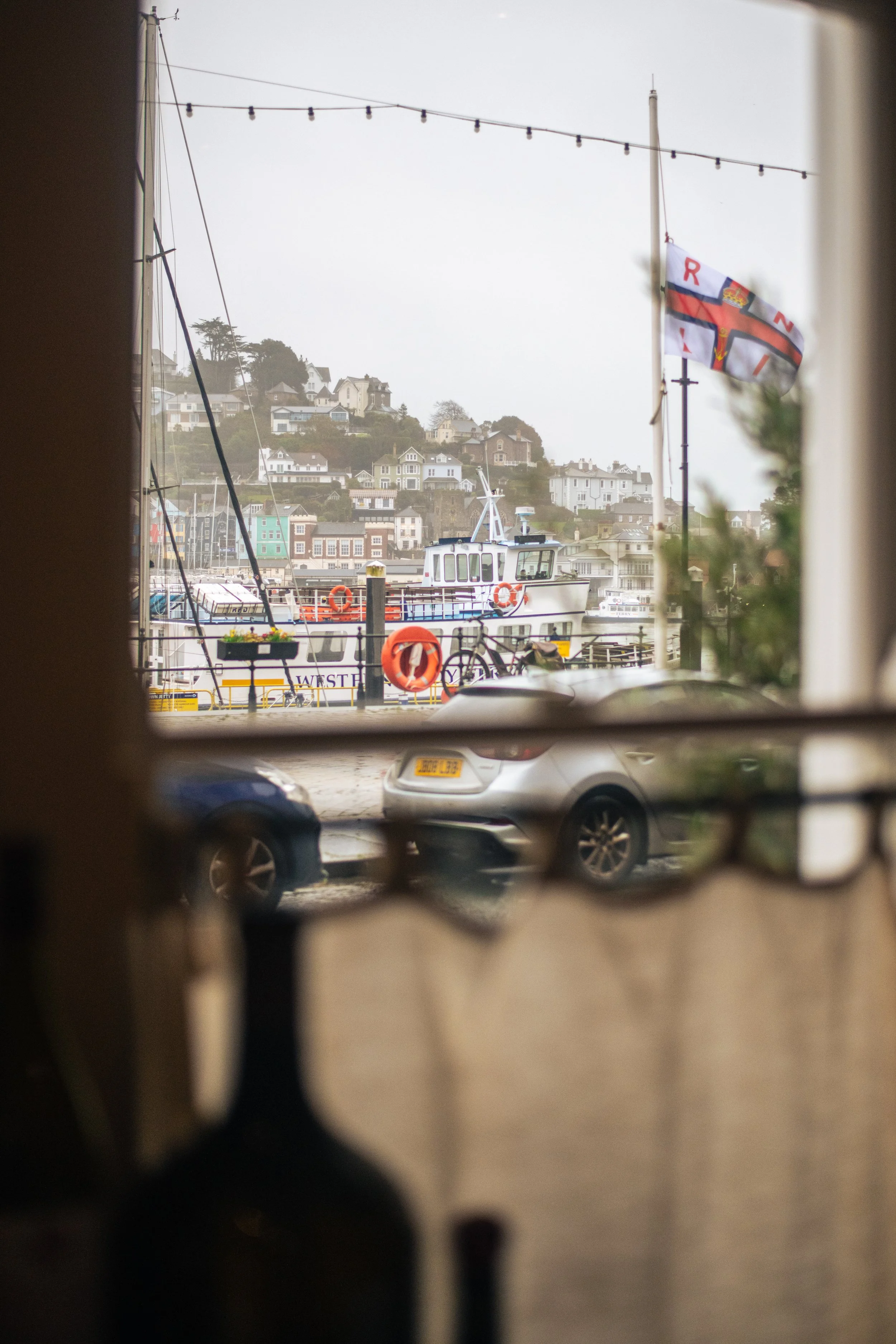 View of a marina with boats, houses on a hillside, and a flag with the Union Jack design, seen through a perforated barrier or window.