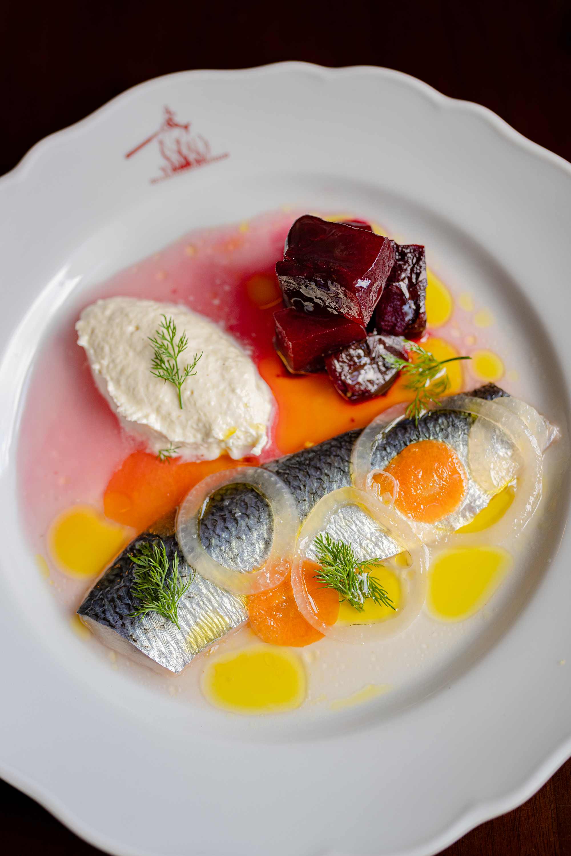 Close-up of a dish with chunks of dark red beets, poached eggs with runny yolks, sliced onions, fresh herbs, and a colorful soup with oil drizzle, in a white bowl with a scalloped edge.