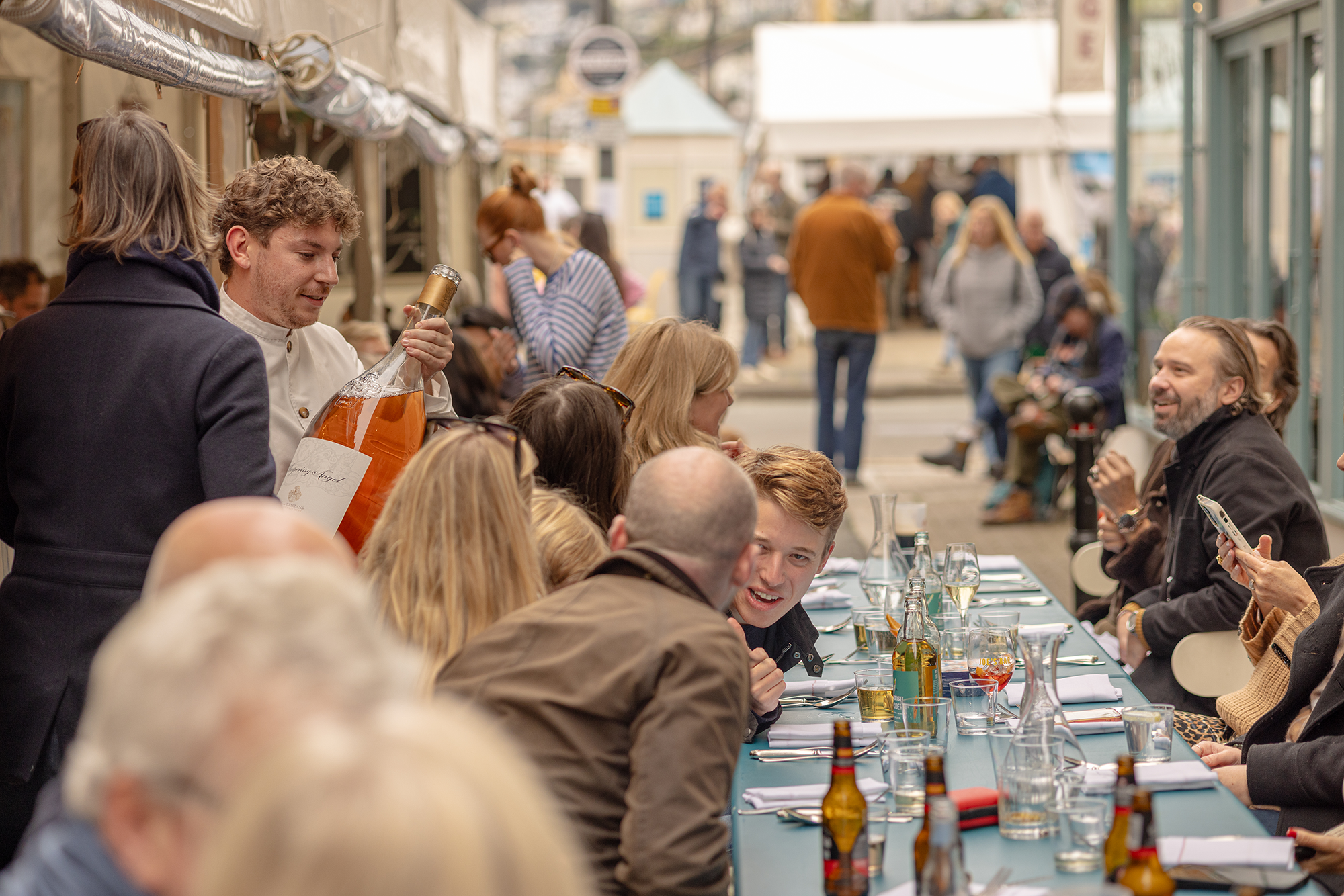People gathered at a long table outdoors, enjoying a meal and drinks in a lively street setting with several onlookers and vendors.