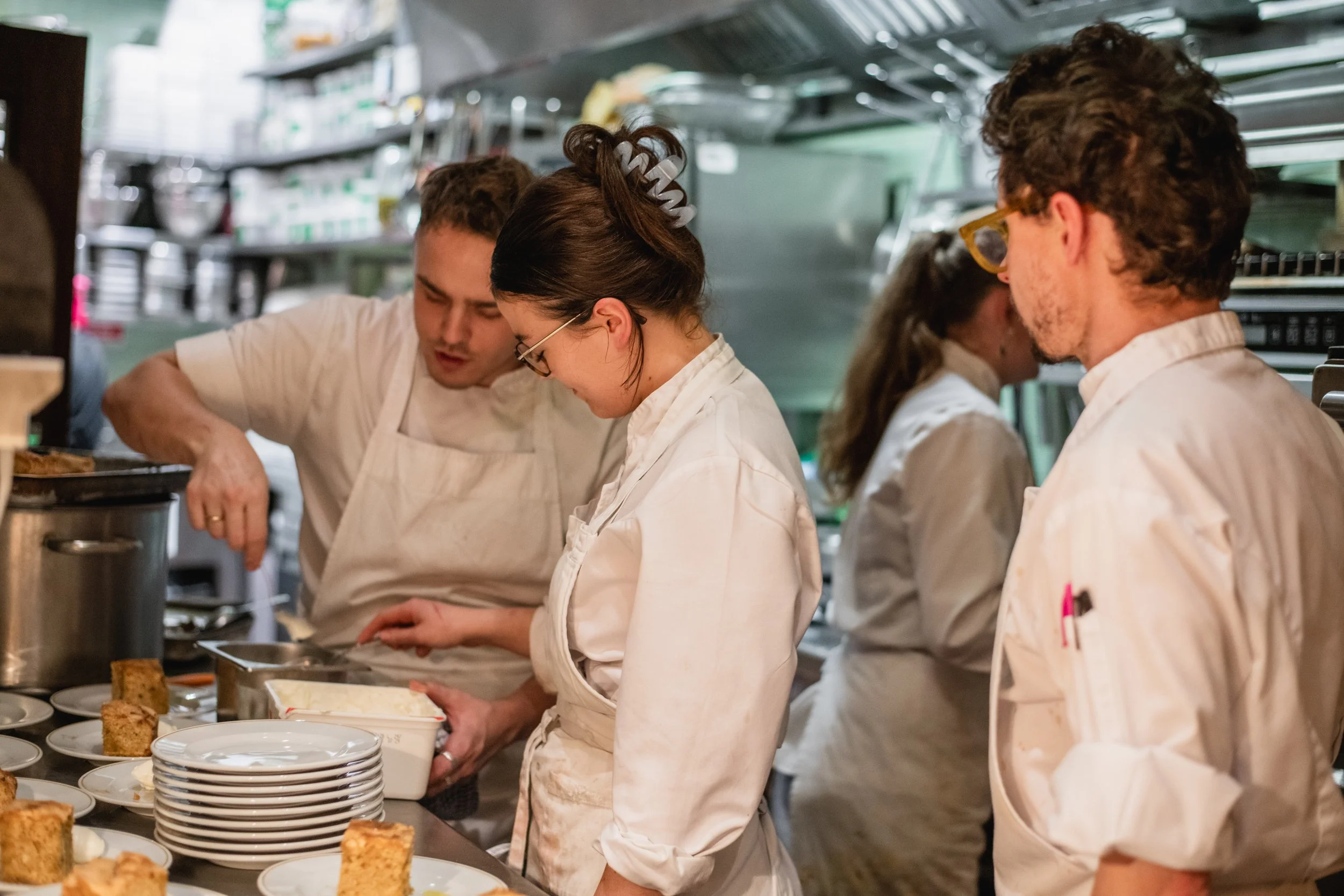 A group of chefs in a kitchen preparing and serving food, with plates and baked goods on the counter.
