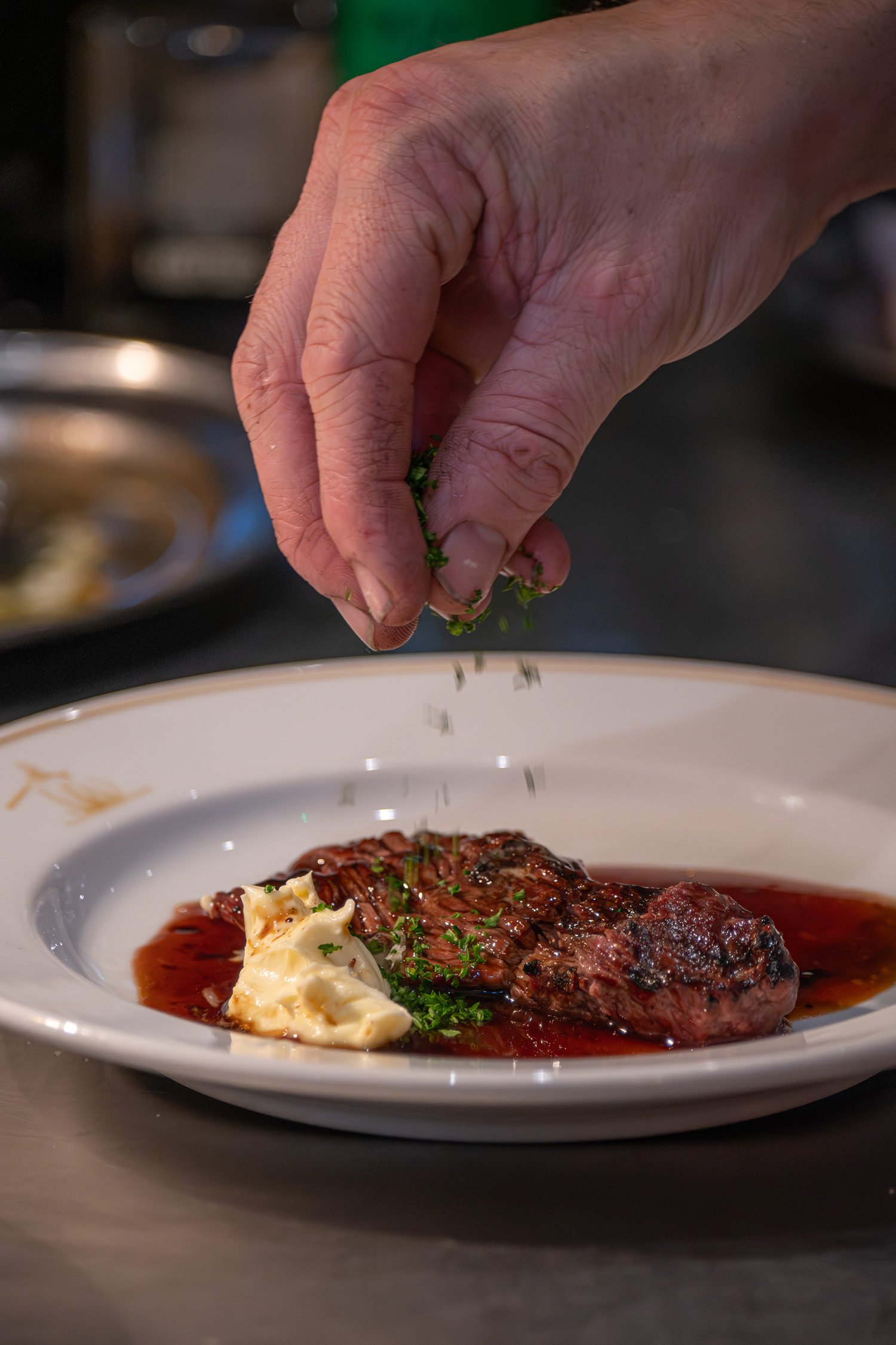 A hand sprinkling chopped green herbs on a plated cooked steak with sauce, butter, and garnish.