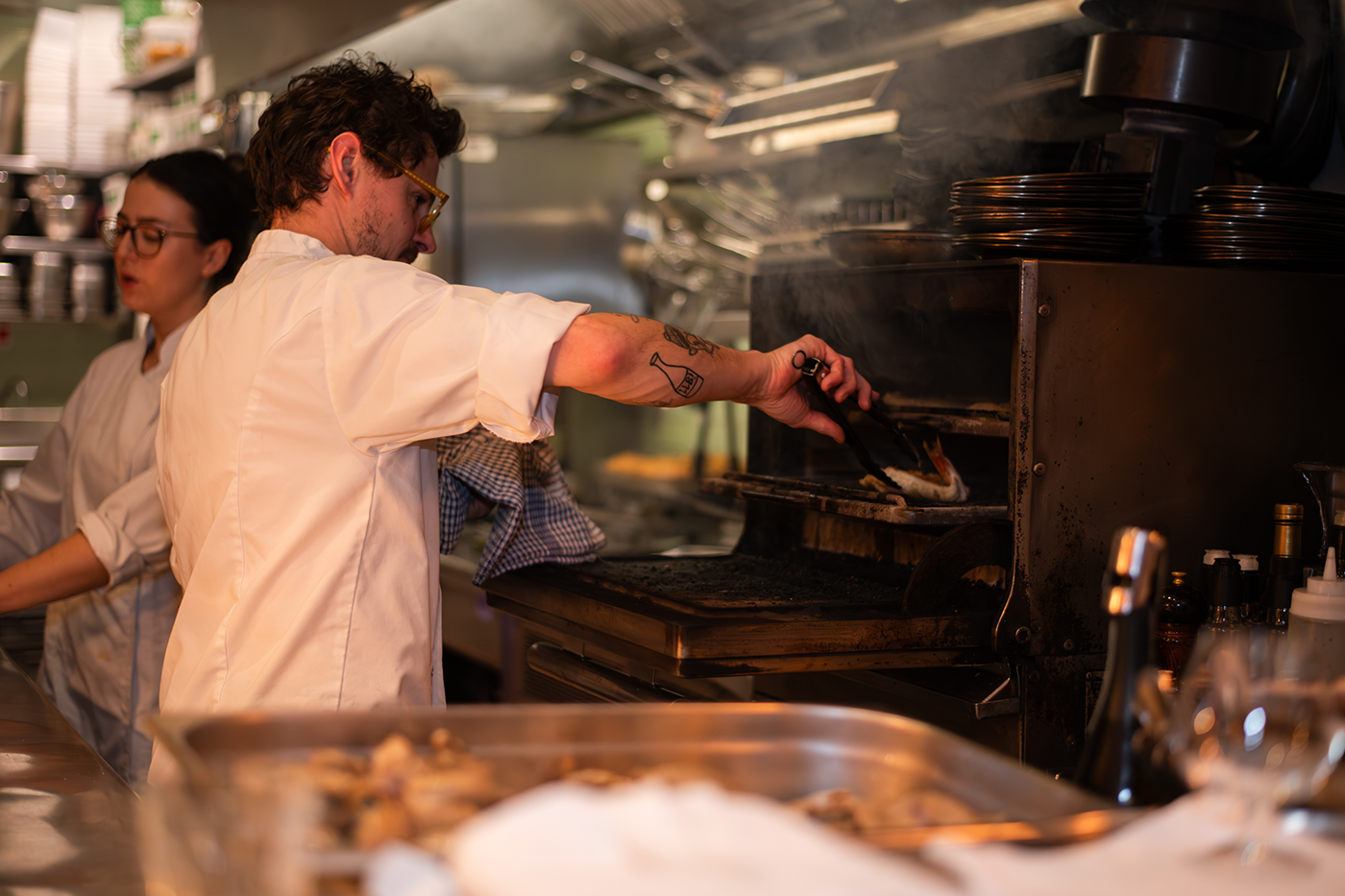 Chef placing a pizza into an oven while another person prepares food in the kitchen.