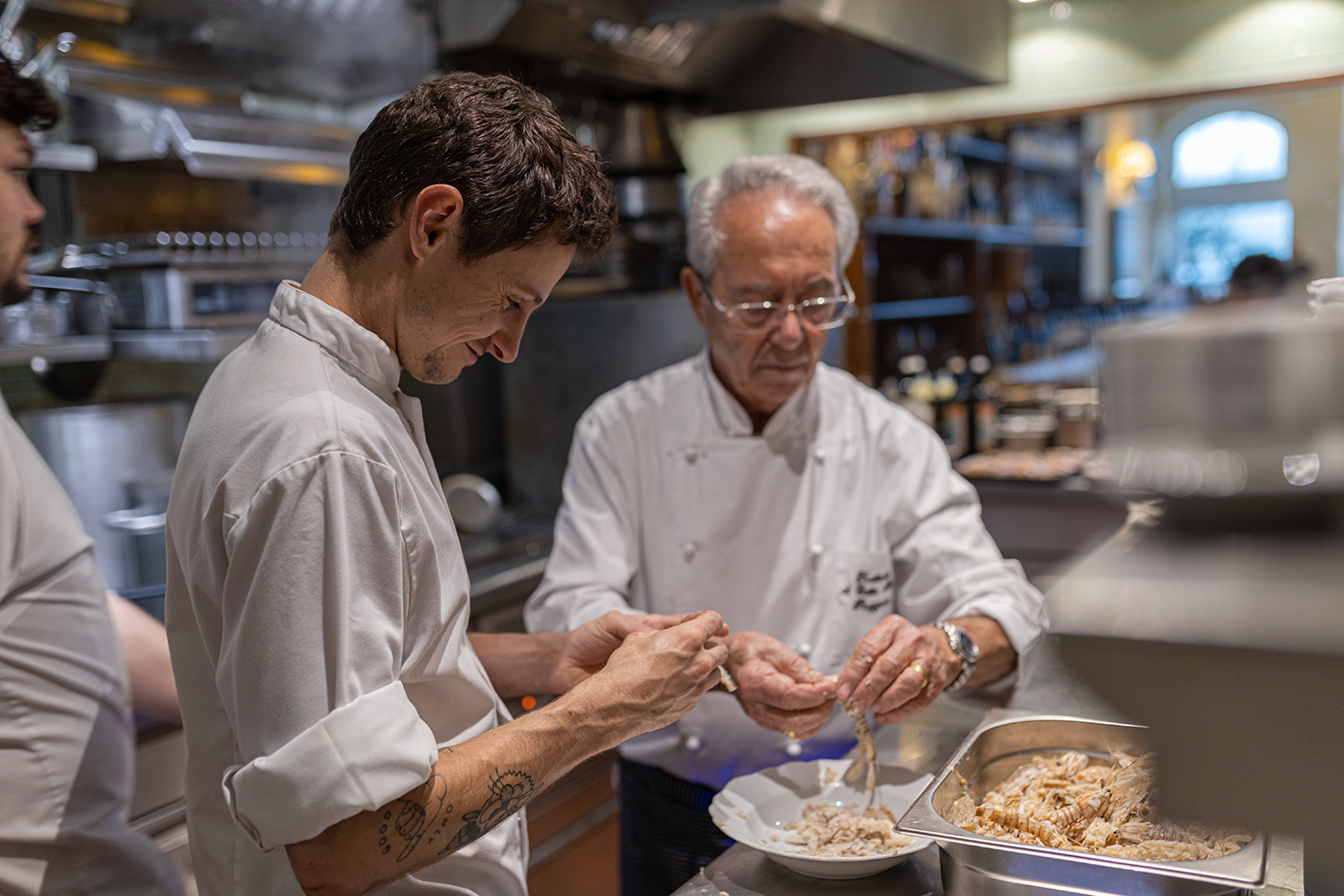 Two chefs, one younger and one older, preparing seafood in a restaurant kitchen, with stainless steel appliances and shelves in the background.