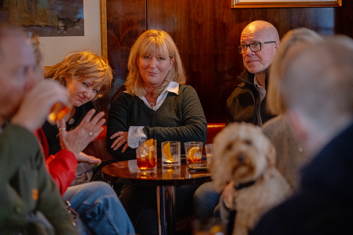 Group of people sitting around a table in a cozy pub or restaurant, with drinks and a dog present