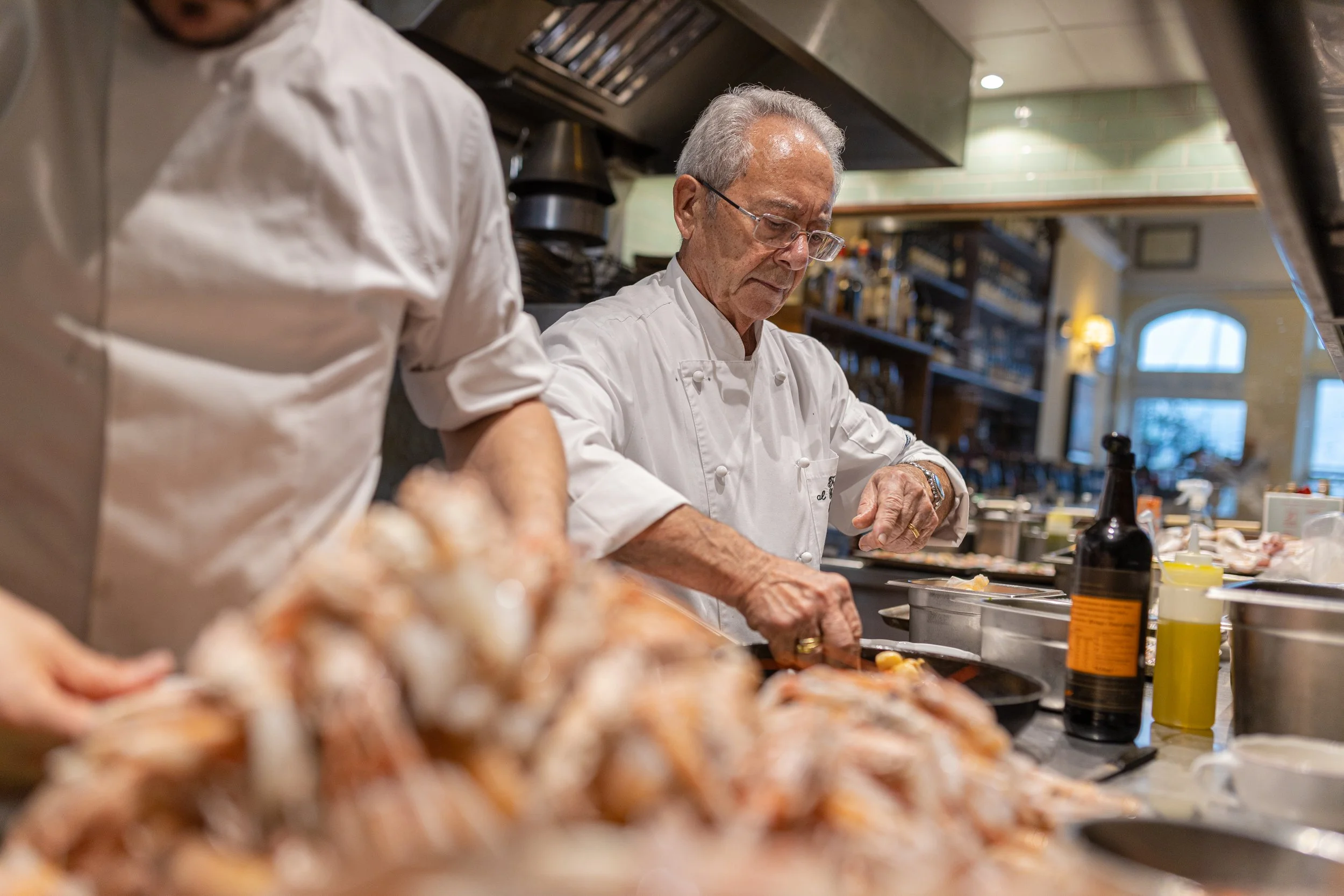 Chef with glasses and white uniform working in a busy kitchen. Foreground shows raw meat being prepared and kitchen utensils and ingredients on the counter.