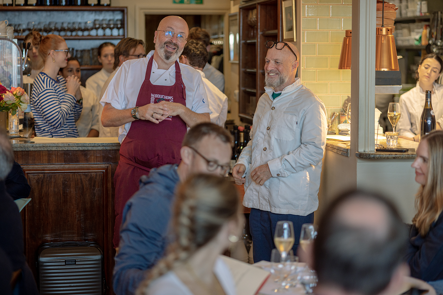 Two chefs speaking to a group of diners in a restaurant, with some staff and guests visible in the background.