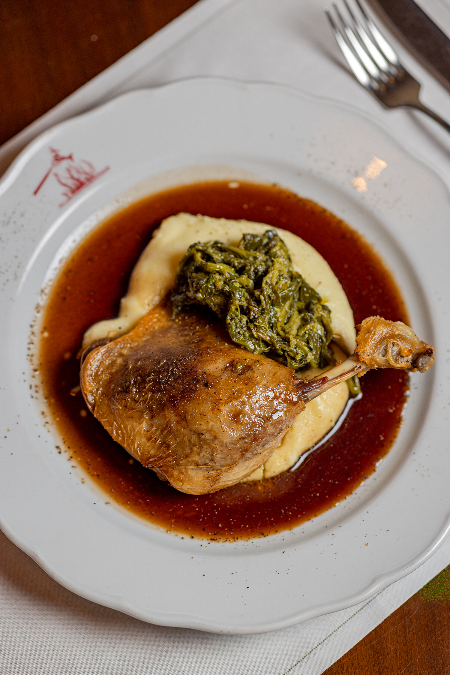 A plate of roasted chicken with mashed potatoes, greens, and brown gravy, served on a white dish with a decorative red emblem, placed on a white placemat with a fork nearby.