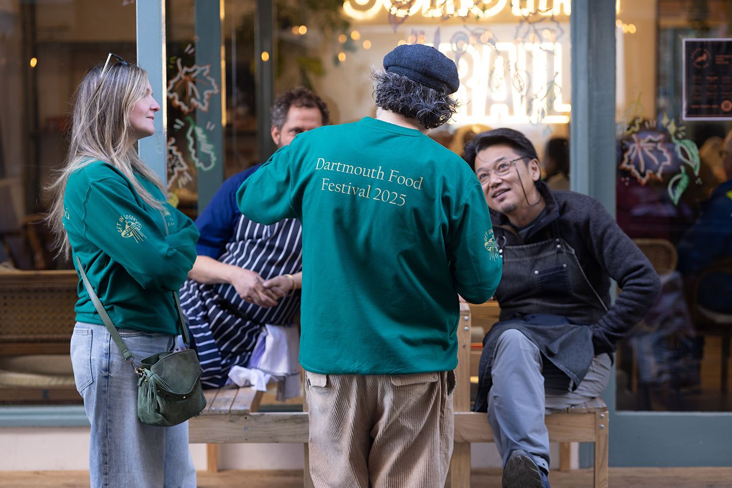 Four people standing and sitting outside a restaurant, engaged in conversation during the Dartmouth Food Festival 2025. One person facing away is wearing a teal jacket with the event name, while others wear casual clothing and aprons.