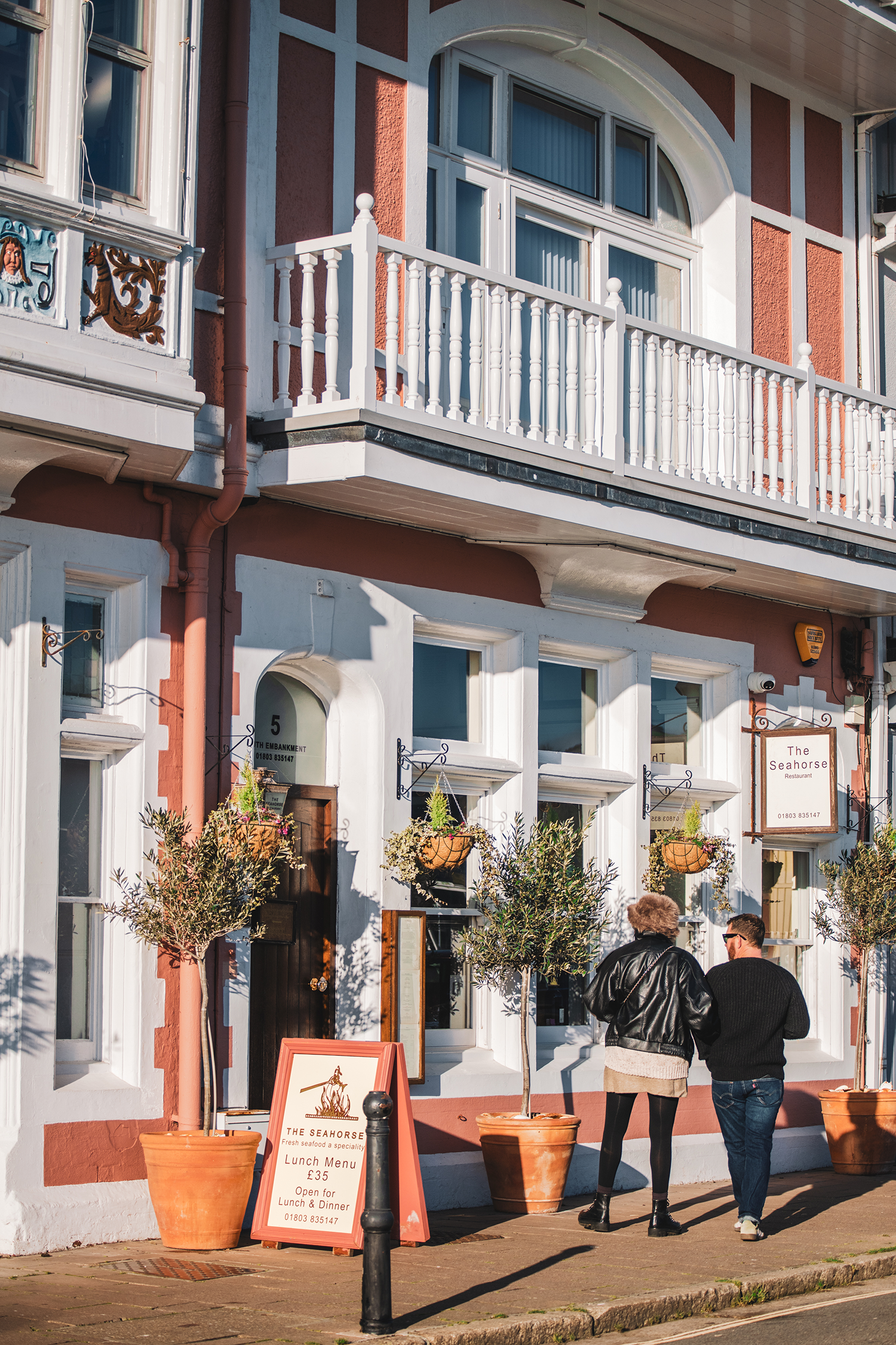 People walking past a restaurant with a signboard outside on a city sidewalk. The building has white-framed windows, a small balcony with a white railing, and decorative details on the facade. Potted plants are arranged along the sidewalk.