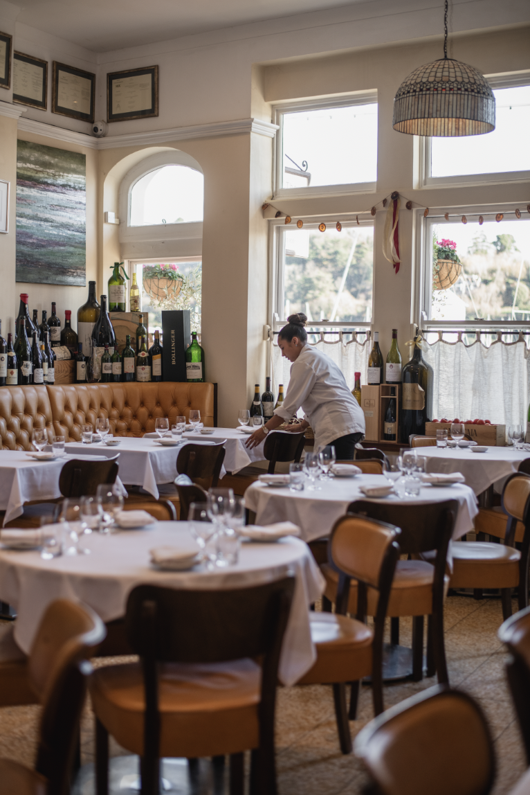A waiter setting tables in a cozy restaurant with white tablecloths, wine bottles on the wall, and large windows letting in natural light.