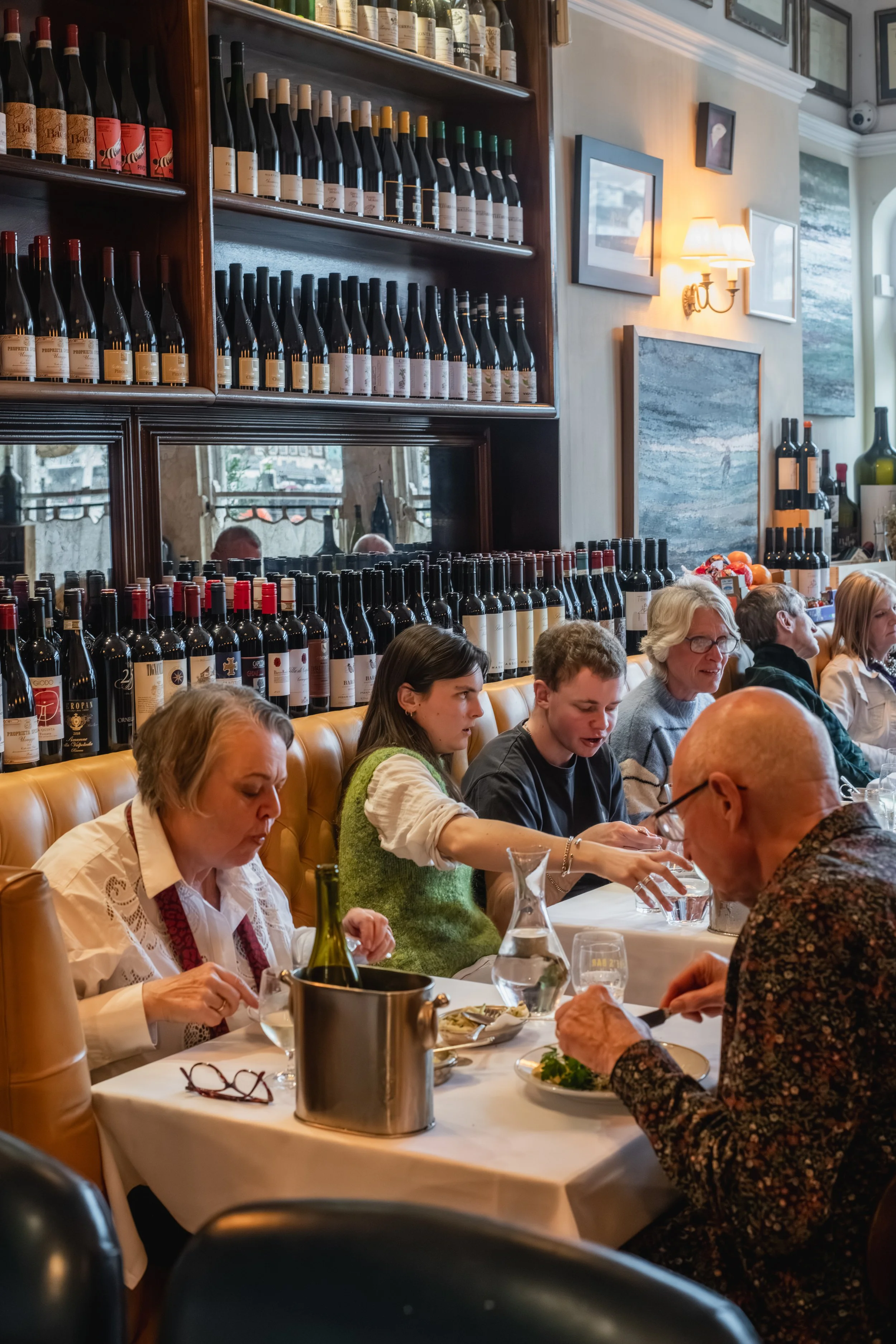 People dining at a restaurant table with wine bottles on shelves in the background.