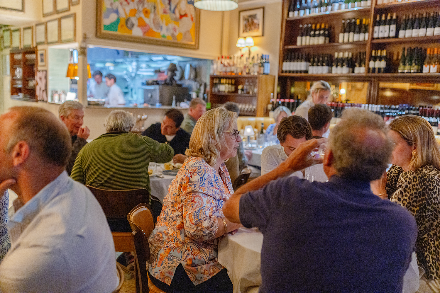 People dining at a restaurant with wine bottles on shelves, a view into the kitchen, and warm lighting.
