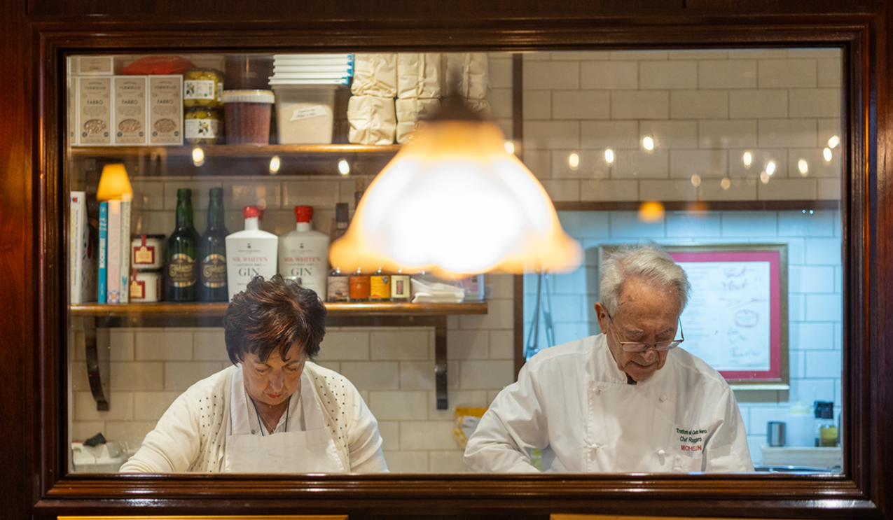 Two chefs working in a restaurant kitchen, viewed through a window. The female chef has short dark hair and is wearing a white apron, while the male chef has gray hair, glasses, and is wearing a white chef's coat. Shelves with bottles, jars, and boxes are visible behind them.