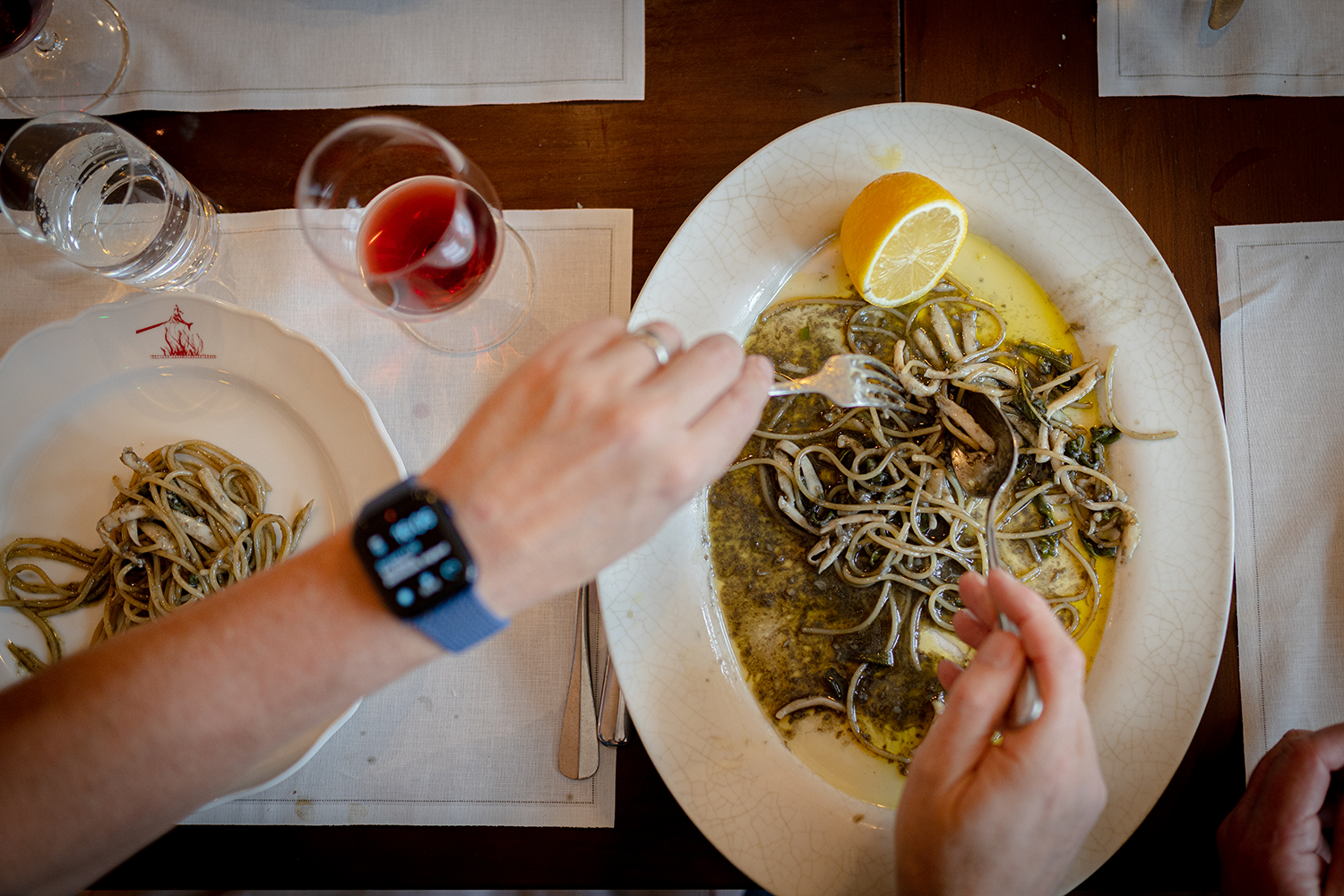 A person with a smartwatch is eating a seafood pasta dish with a lemon wedge at a dining table that has a white placemat and a glass of red wine.