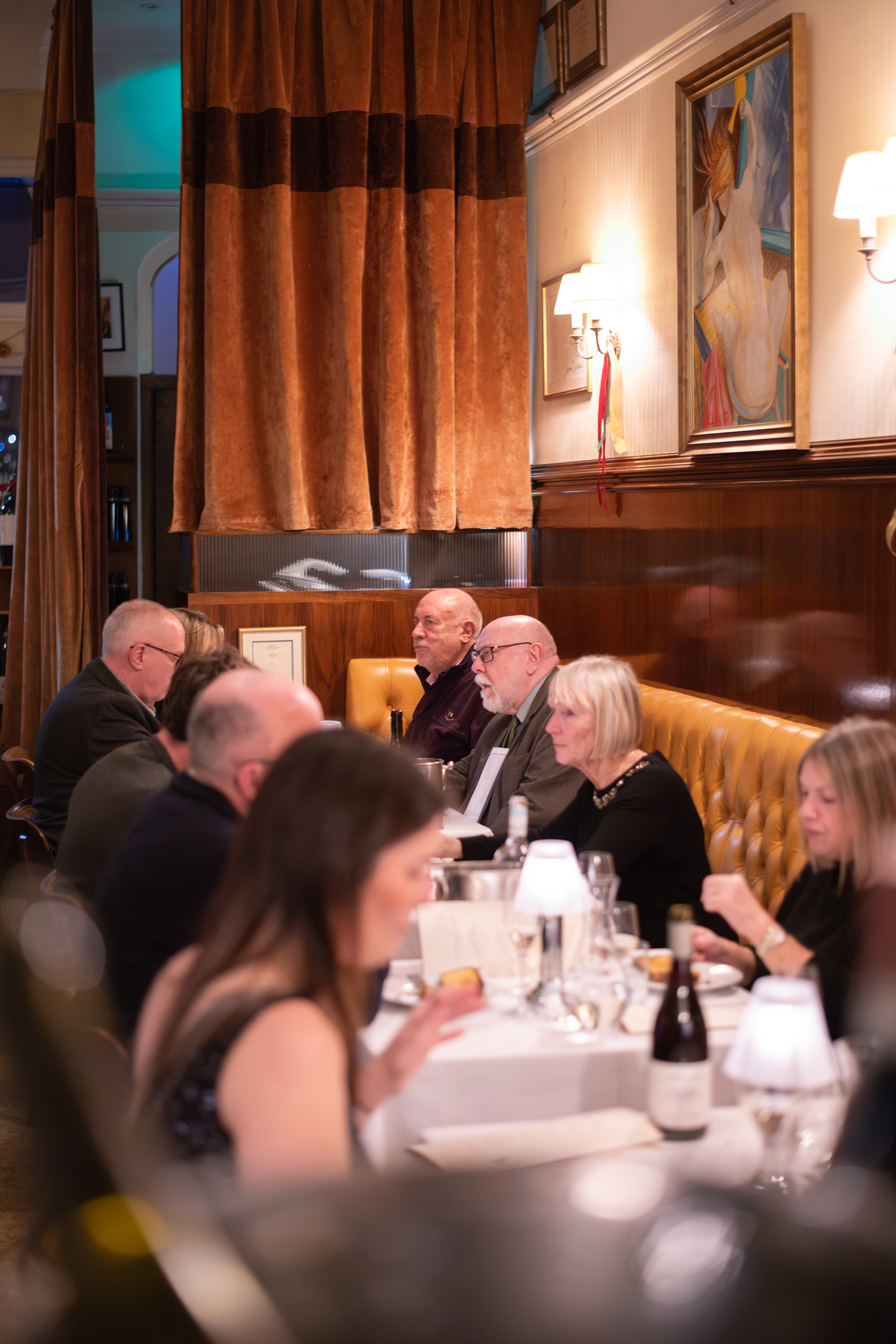 People gathered at a restaurant or banquet for a meal, sitting at a long table with white tablecloths, glasses, bottles, and lamps, in a warmly lit room with brown curtains and framed artwork on the walls.