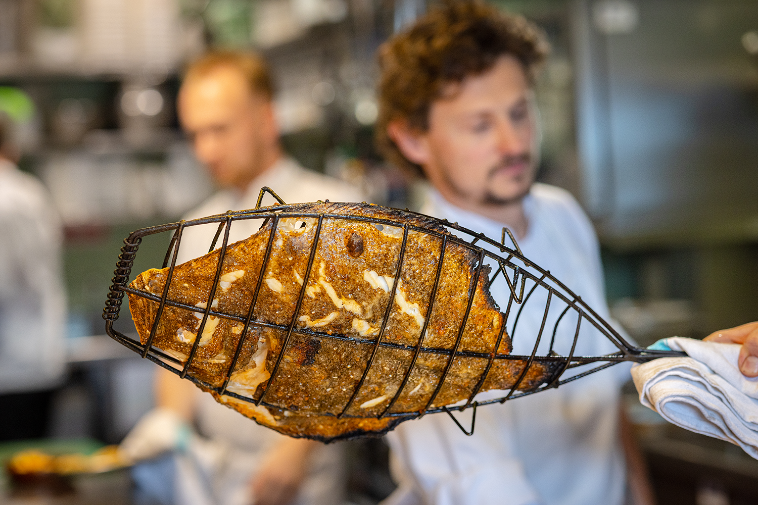 Chef holding a wire basket with a cooked fish in a restaurant kitchen.