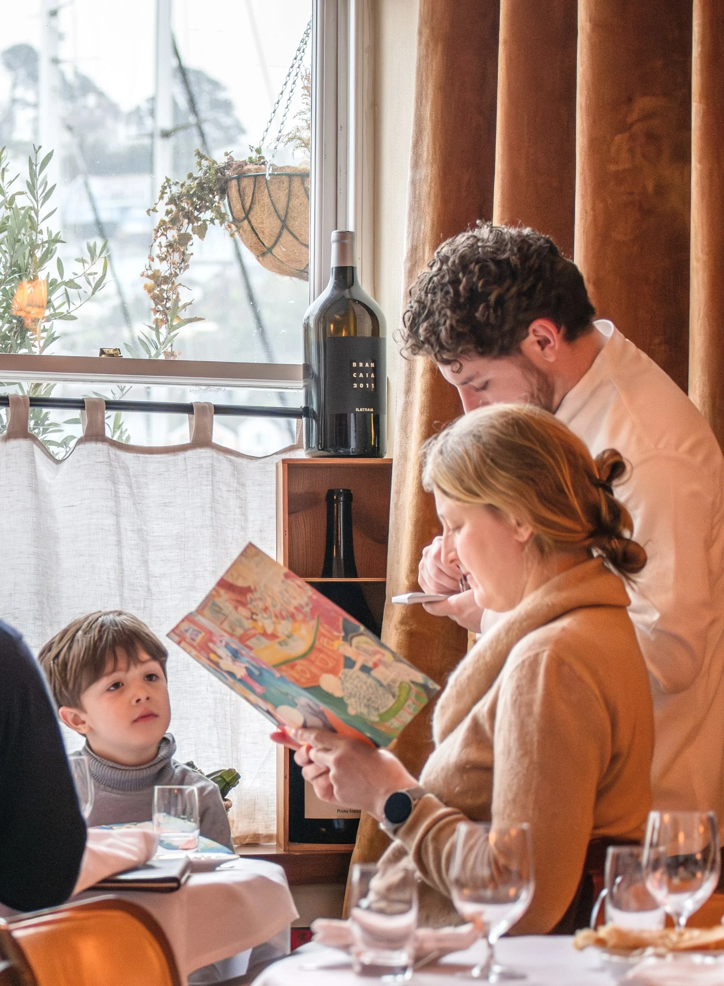 People sitting at a dining table during a meal, with a young boy looking at the woman reading a colorful card or brochure. A window with curtains and a hanging plant can be seen in the background.