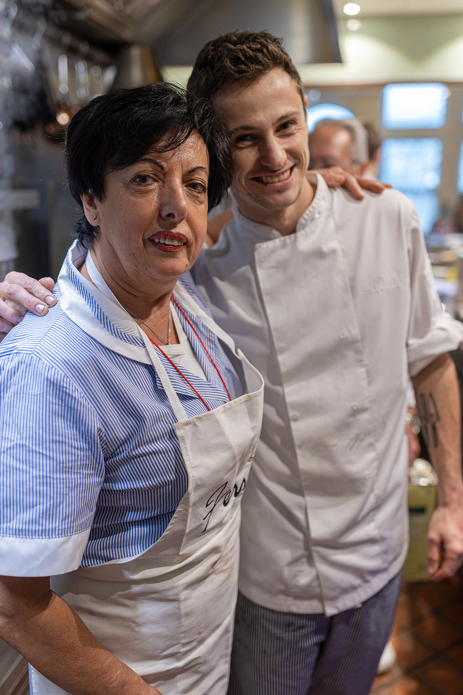 Two smiling chefs in a kitchen, close to each other, wearing aprons, with the woman on the left having short dark hair and the younger man on the right having short curly hair.