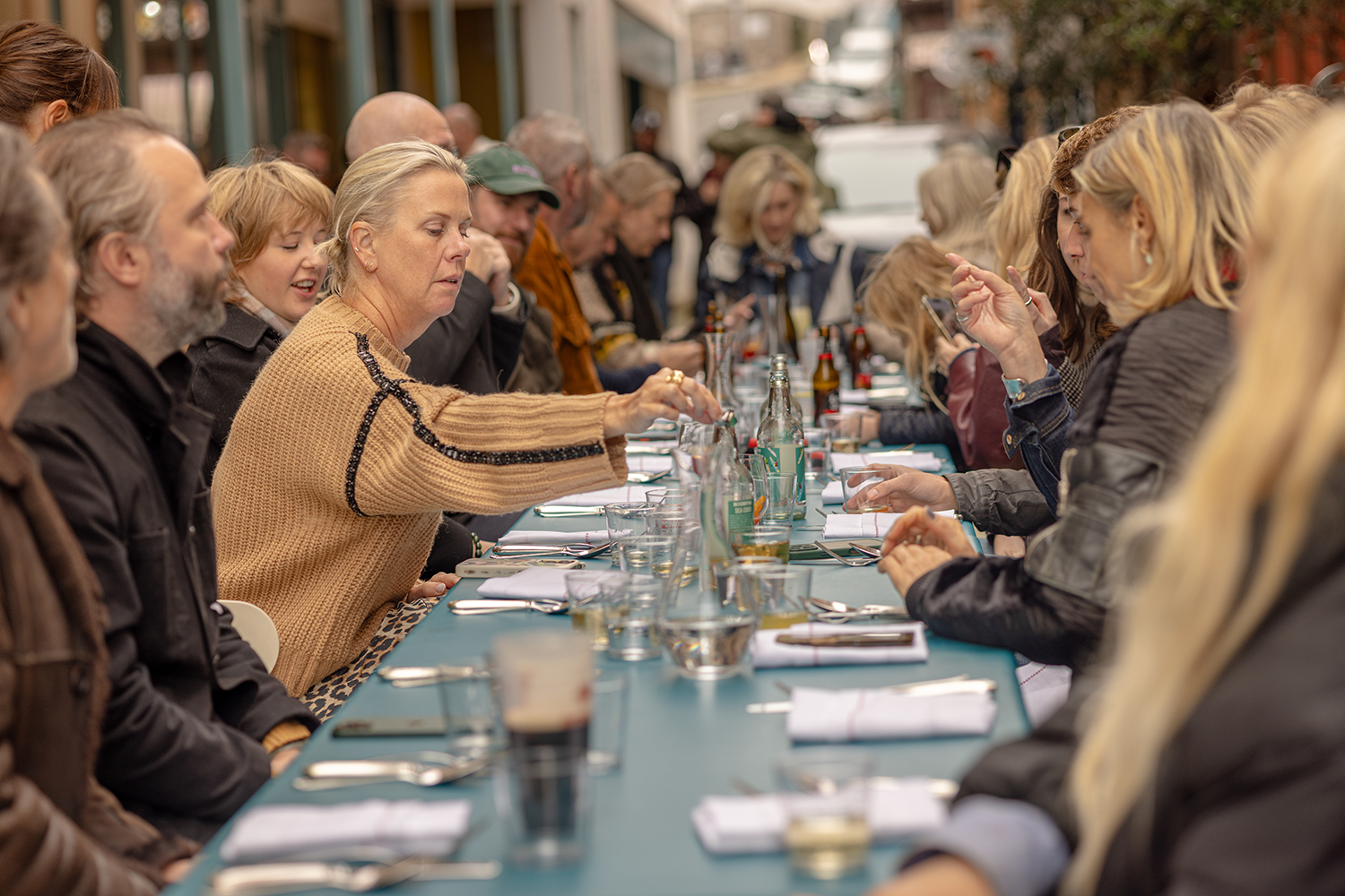 A group of people sitting at a long outdoor table for a meal or gathering, engaged in conversation and using their phones.