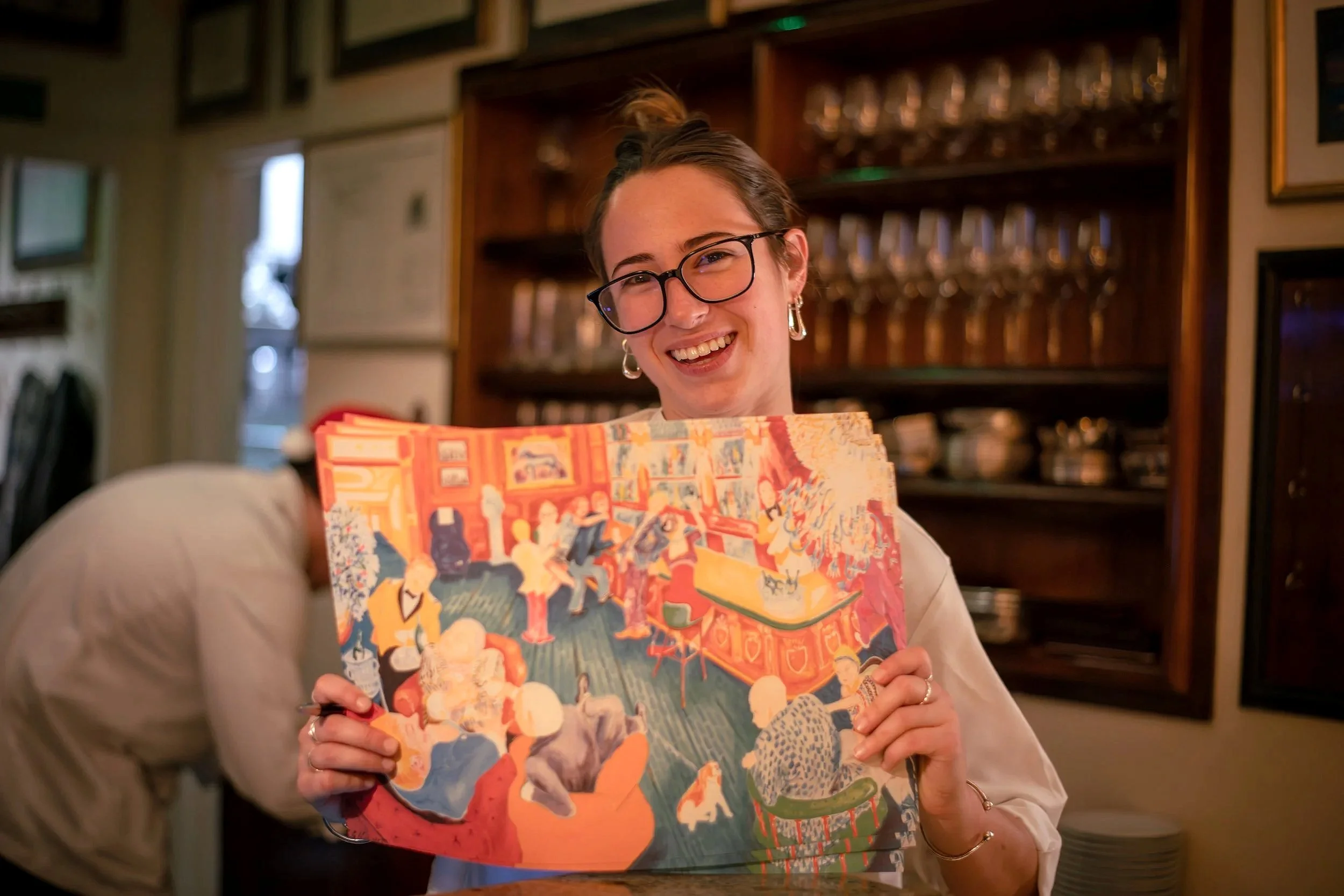 A woman wearing glasses and earrings smiling while holding a colorful illustrated book in a cozy room with framed pictures and a liquor cabinet in the background.