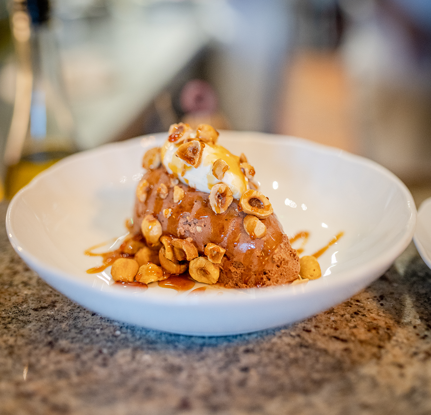 A dessert in a white bowl featuring a scoop of ice cream on top of a brownie, drizzled with caramel sauce, chopped nuts, and a dollop of whipped cream.