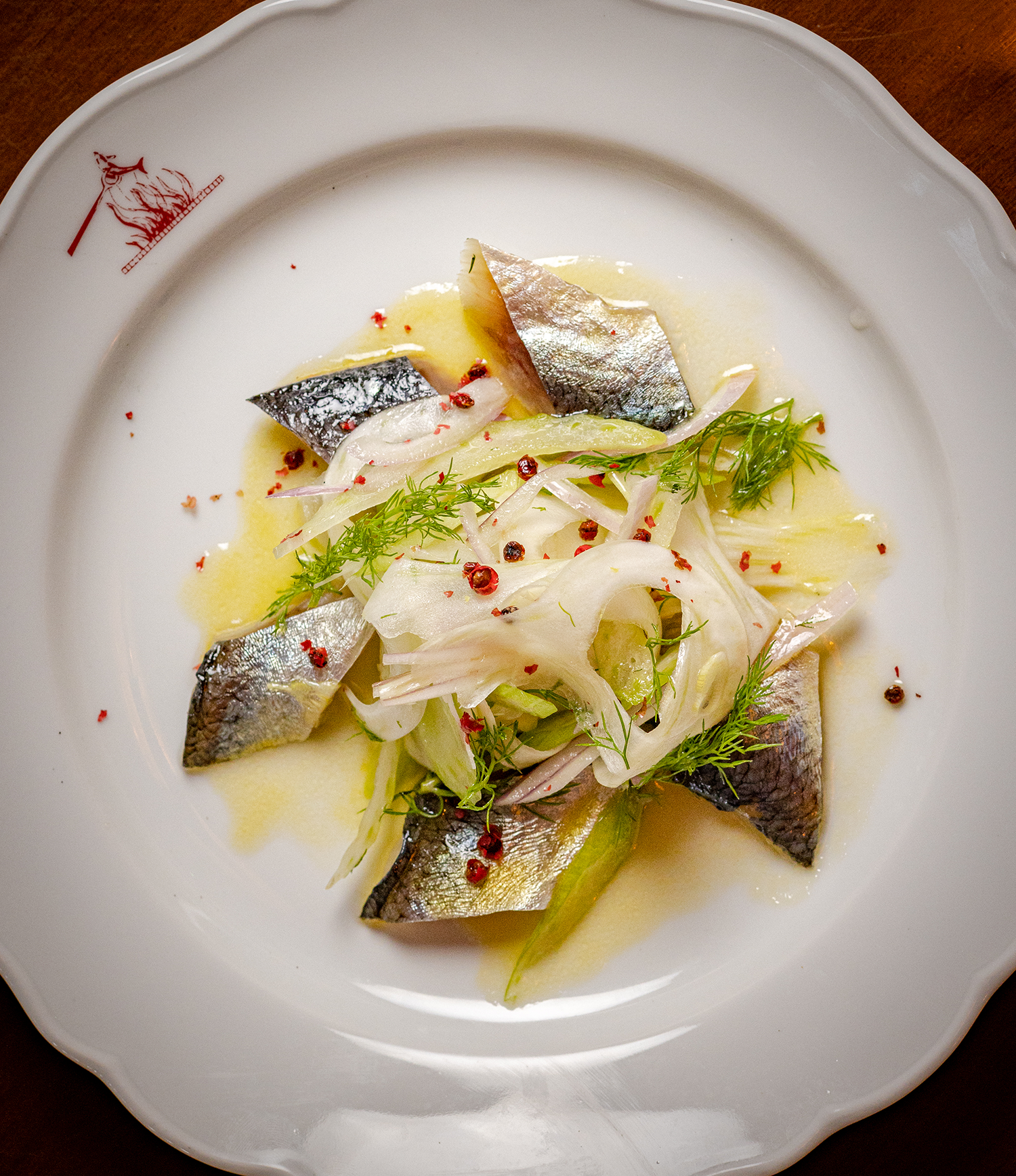A white plate with a decorative red design on the edge, holding pieces of fish with silvery skin, served with sliced onions, fresh herbs, and a light yellow sauce with red pepper flakes.