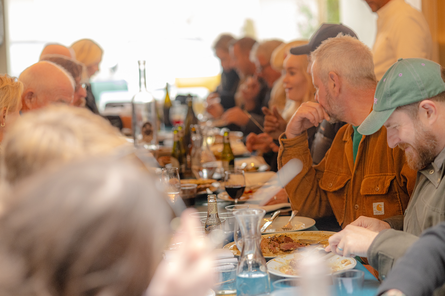 People sitting at a long dining table enjoying a meal, with food, drinks, and bottles on the table in a warmly lit room.