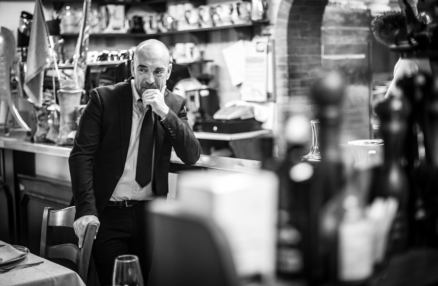 A man in a suit and tie standing in a restaurant or cafe, resting his hand on a chair, with shelves, bottles, and flags in the background.