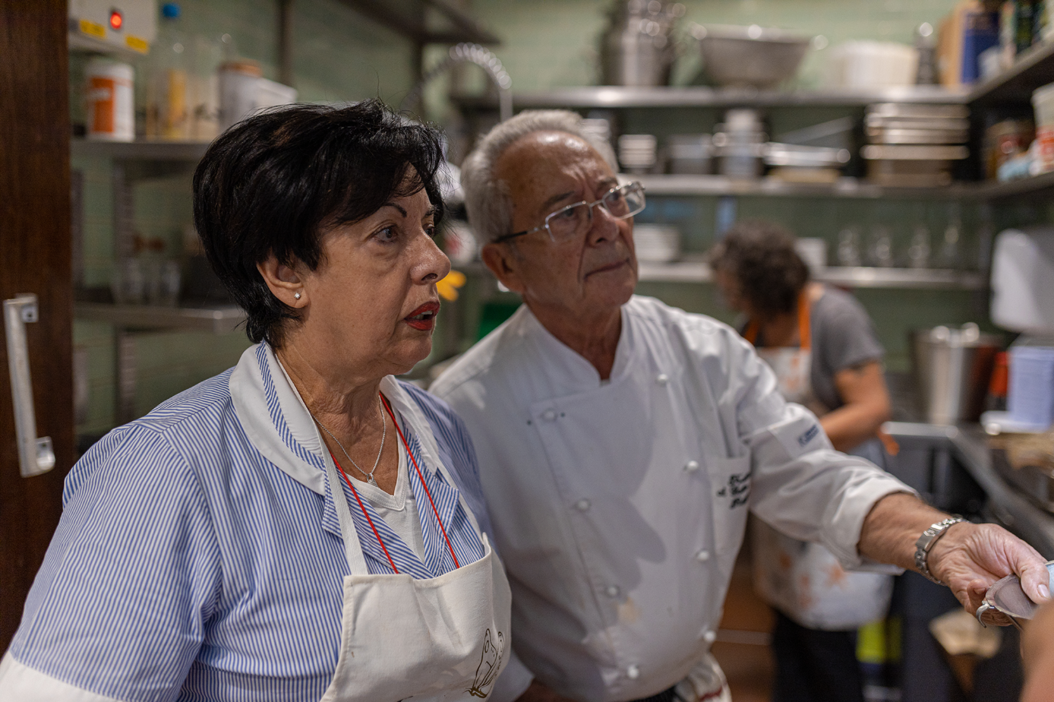 Two elderly individuals, a woman and a man, wearing aprons in a professional kitchen, engaged in a culinary activity, with shelves of kitchen supplies and another person in the background.