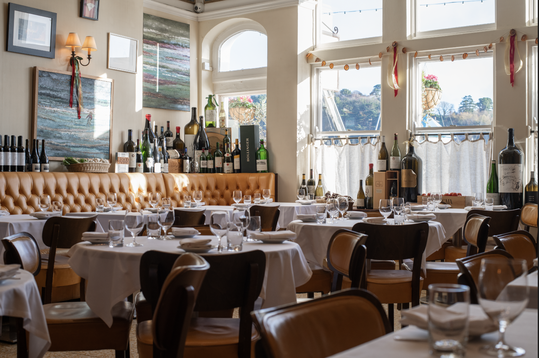 A decorated restaurant dining area with white tablecloths, glasses, and cutlery set for diners. The room has large windows letting in natural light, with some wine bottles and decorative items on a shelf along the wall, and outdoor views of trees and hills.