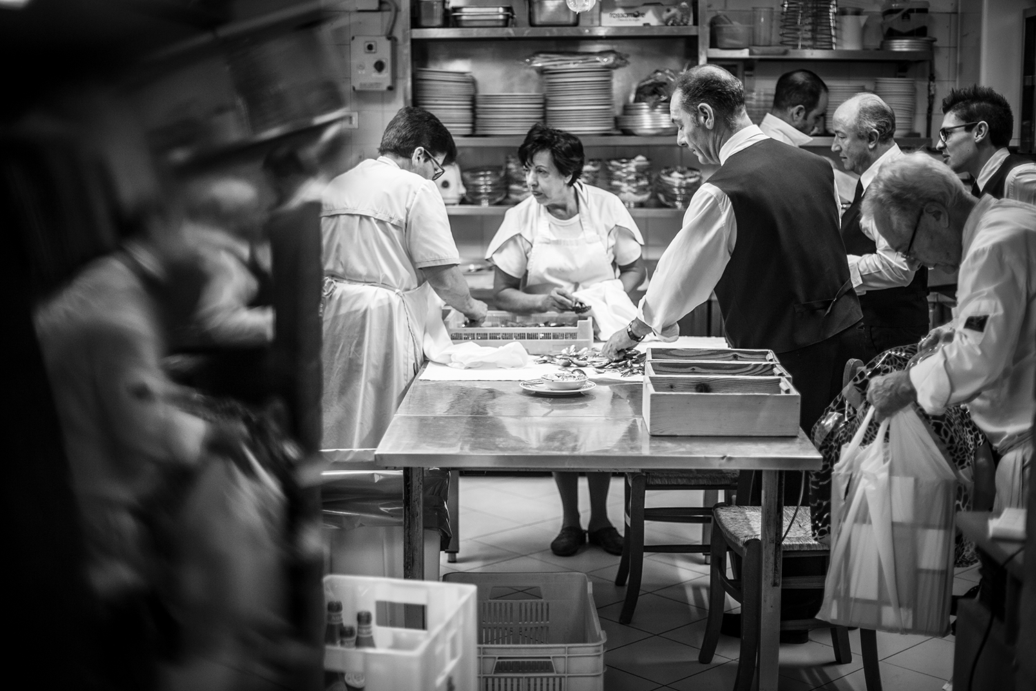 Black and white photo of a busy kitchen with people preparing food. Several chefs and staff are gathered around a table, working with their hands. Shelves with dishes are visible in the background.