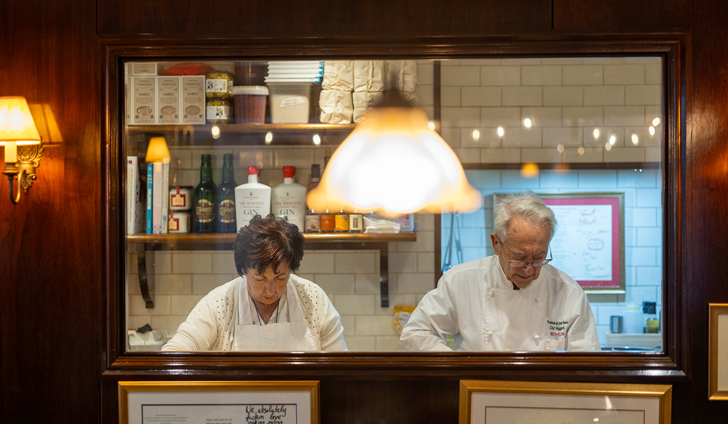 Two chefs working in a kitchen seen through a window in a dining room, with warm lighting and framed documents below.