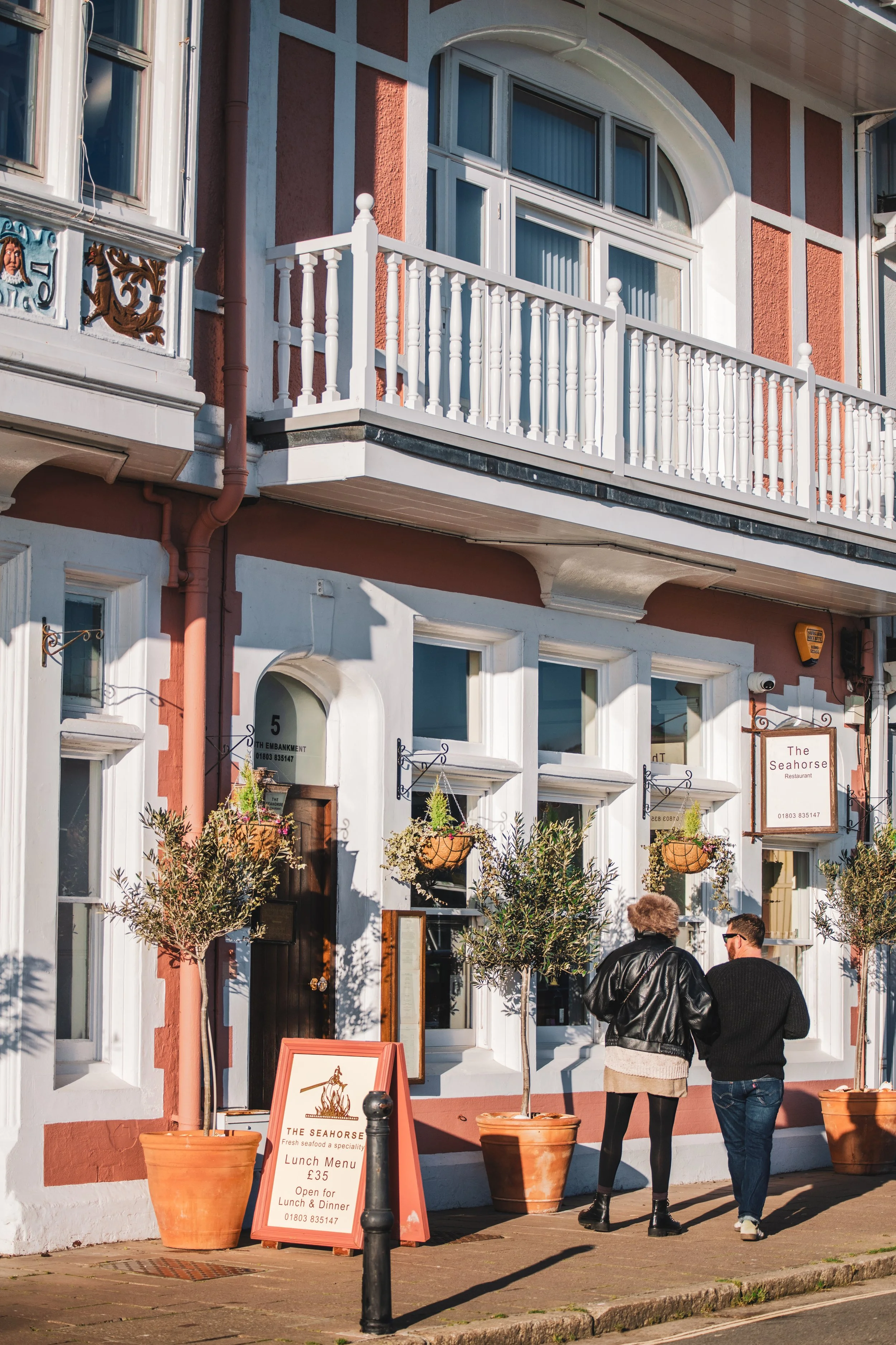 Two people walking past a restaurant called The Seahorse, which has a pink and white exterior with potted plants and a lunch menu sign outside.