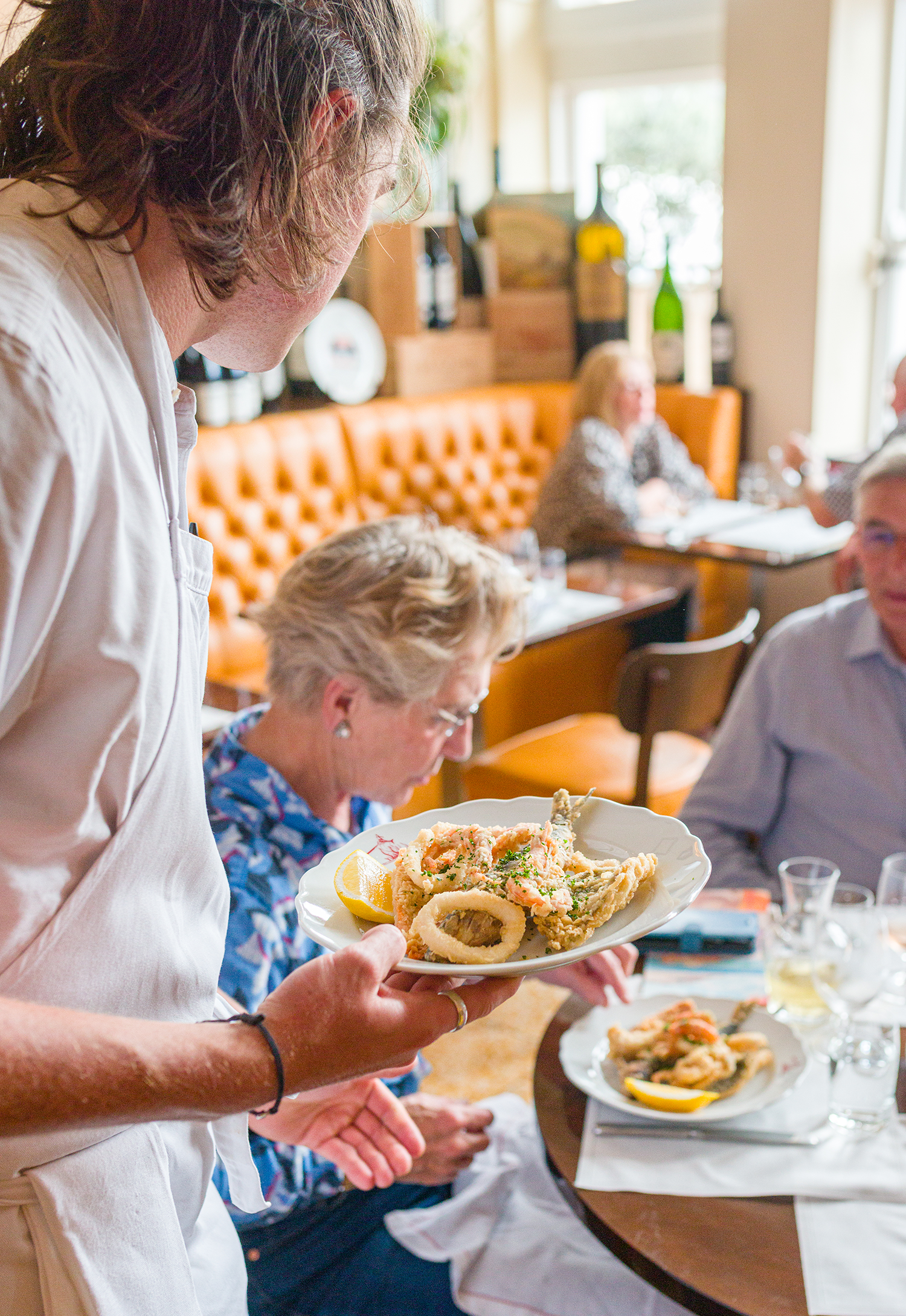 A woman in a white apron holding a plate of fried seafood, including calamari and onion rings, at a restaurant or gathering table with other people and food in the background.