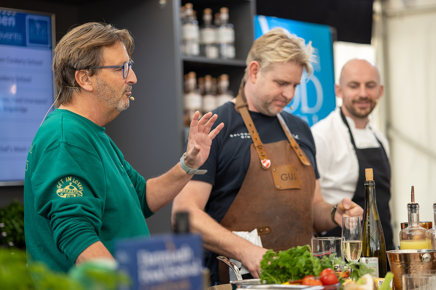 Three men engaging in a cooking demonstration or class, with one man speaking and gesturing, while the others listen, surrounded by ingredients and cooking utensils.