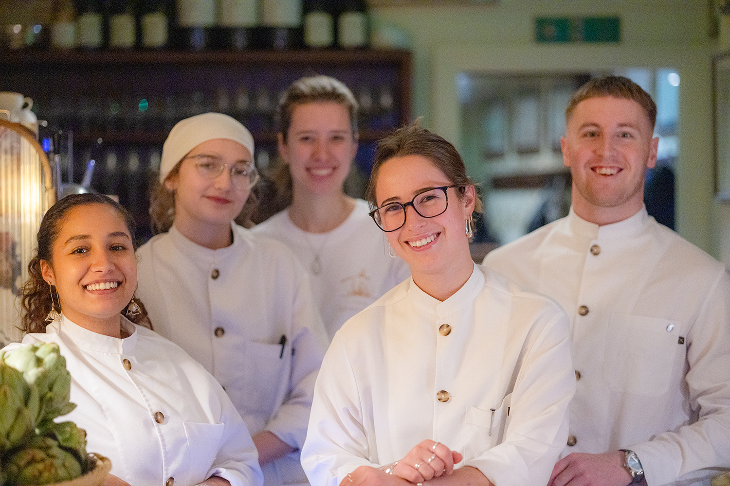 Group of five chefs, four women and one man, smiling in a restaurant kitchen, wearing white chef uniforms.