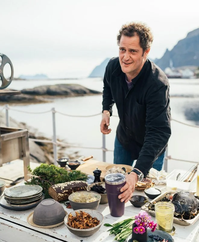 Man outdoors at a table with various dishes and drinks near a rocky coastline and mountains.