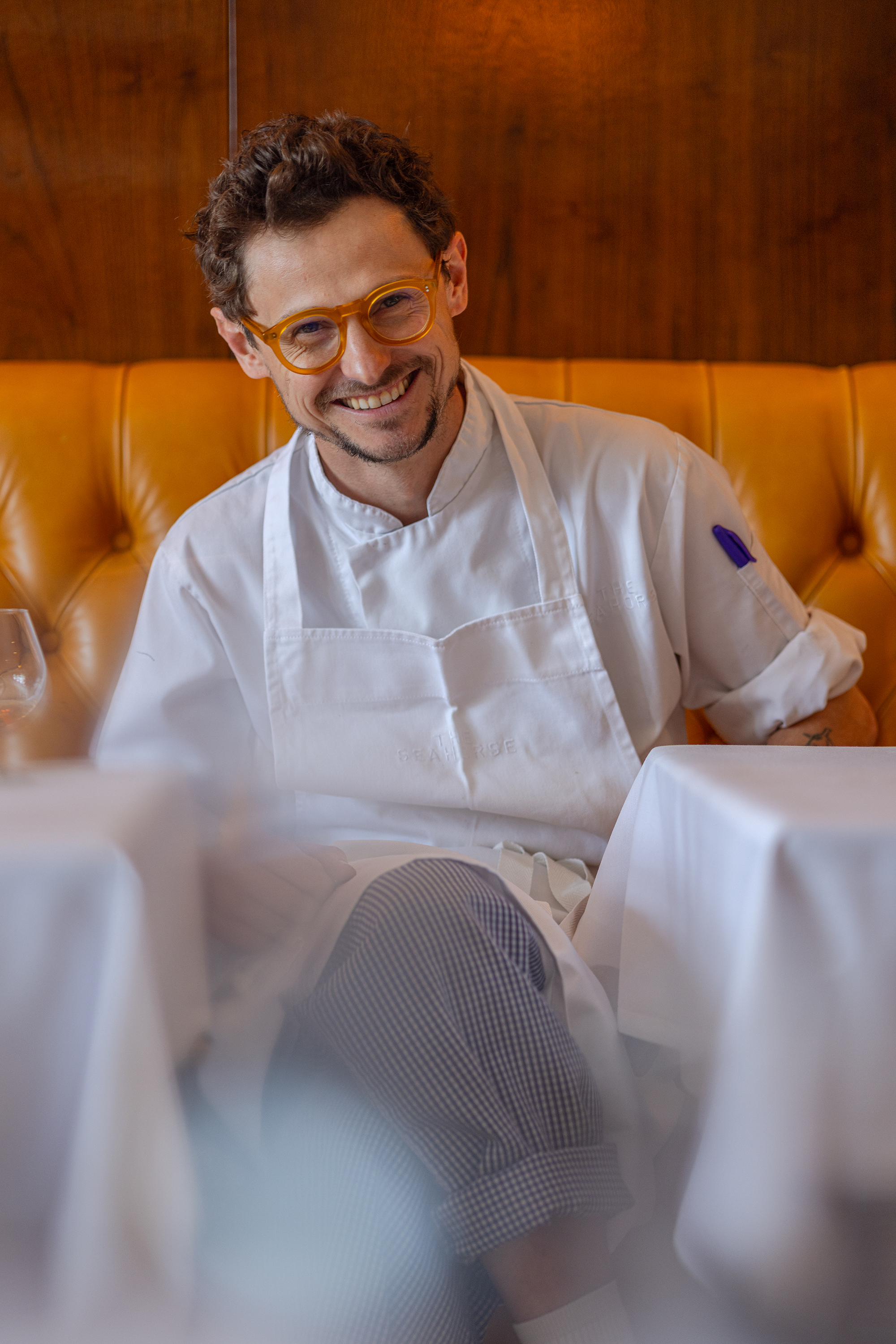 Smiling man with curly brown hair, glasses, and a mustache, wearing a white chef's coat, sitting at a restaurant table with white tablecloth, wooden panel wall background.